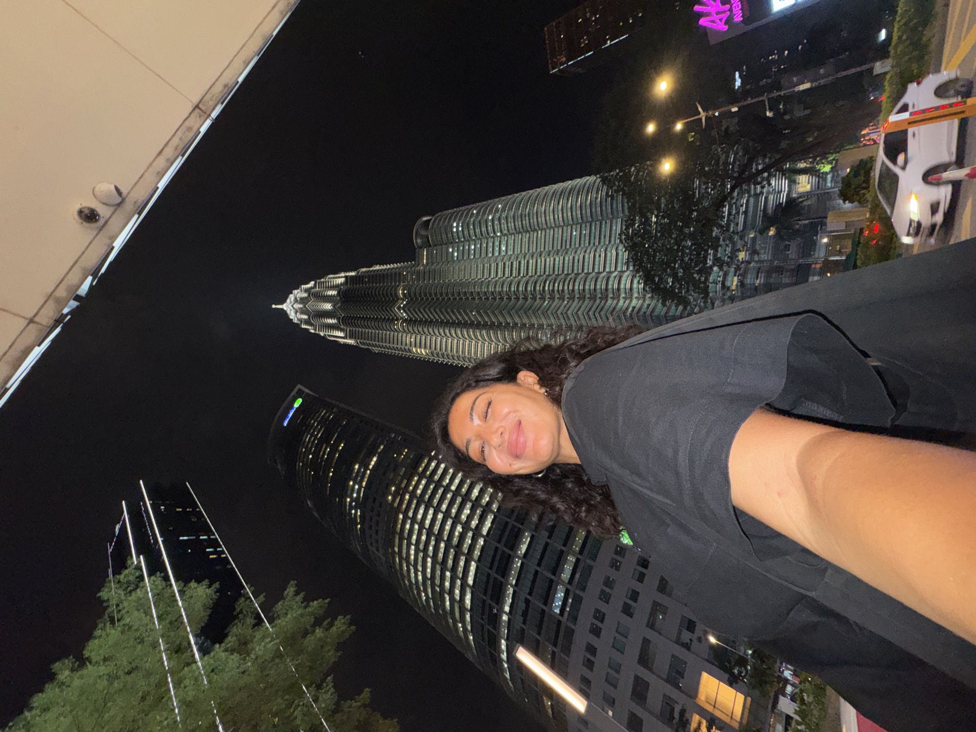 A young woman is captured in a selfie-style photograph at night, showcasing the illuminated cityscape of Kuala Lumpur, Malaysia. She is positioned in the foreground, with her head slightly tilted, sporting long, dark, curly hair and a broad smile with her tongue playfully extended. She wears a dark top.
The background is dominated by iconic skyscrapers, most notably the towering, illuminated Petronas Twin Towers in the center-left. To the right of the woman, another tall, modern building with many lit windows stands prominently. Further into the background on the far left, another high-rise building with vertical strip lighting is visible.
The scene is set at night, with the dark sky contrasting sharply with the bright artificial lights of the city. Streetlights, glowing windows, and architectural lighting create a vibrant nocturnal atmosphere. A white car, appearing somewhat blurred as if in motion, is visible on the right, alongside some trees. An overhead architectural element, possibly part of a building facade or bridge, with what appears to be security cameras, can be seen in the upper left. A visible sign on a building in the mid-ground displays the text "AVENUE K" in bright pink/purple. The image conveys a sense of excitement and enjoyment amidst a bustling urban environment.