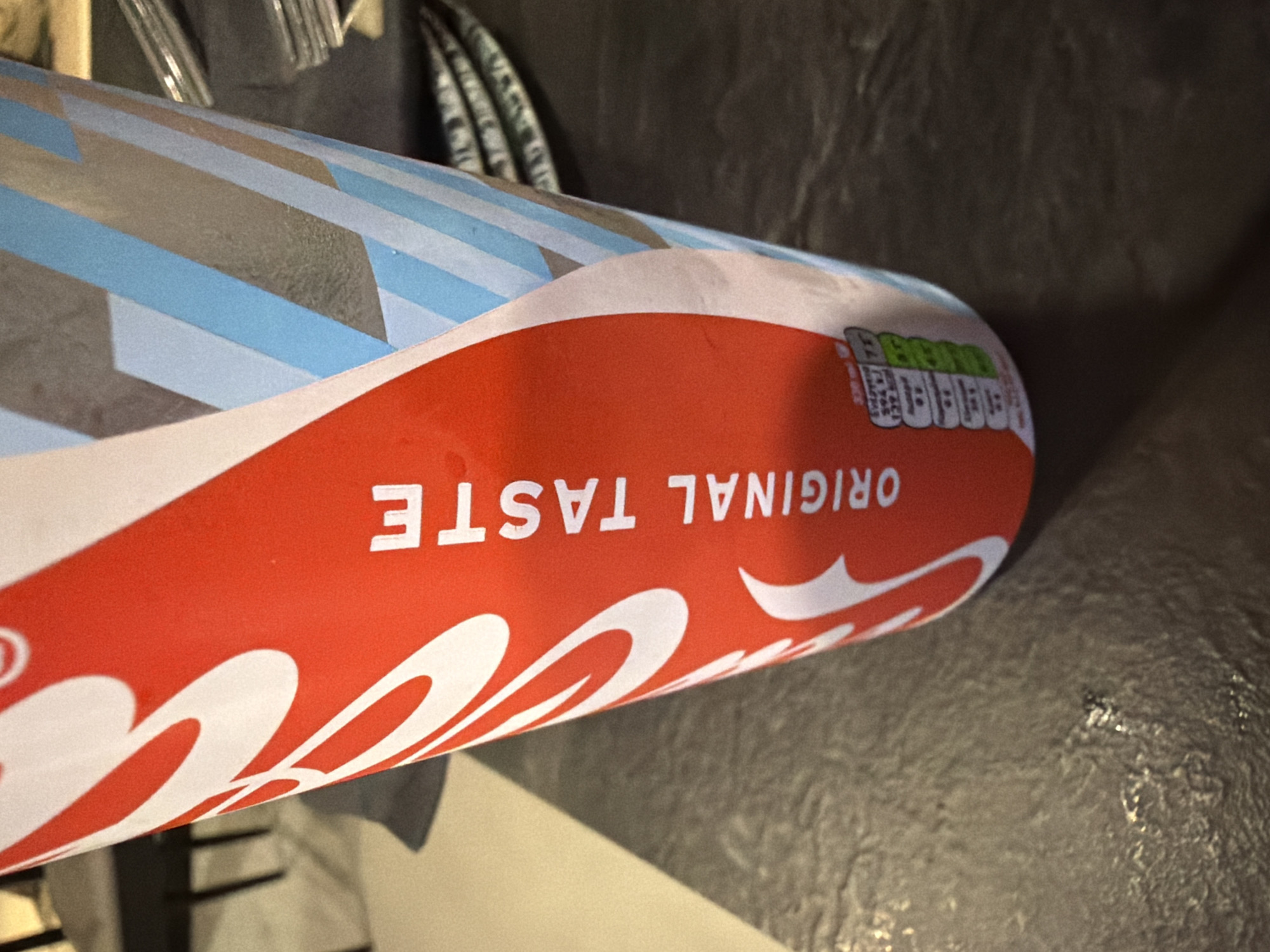 A close-up, low-angle shot focuses on a red and white Coca-Cola cup with a portion of blue and grey geometric patterns on the right side. The text "ORIGINAL TASTE" is vertically printed in white on the red portion of the cup. A small nutrition label with green and red bars is visible at the bottom of the cup. The cup is placed on a dark, textured surface, possibly a table. In the background, out of focus, are dark chair legs and a stack of metallic cutlery. The lighting suggests an indoor setting. The location is Kiti, Cyprus.