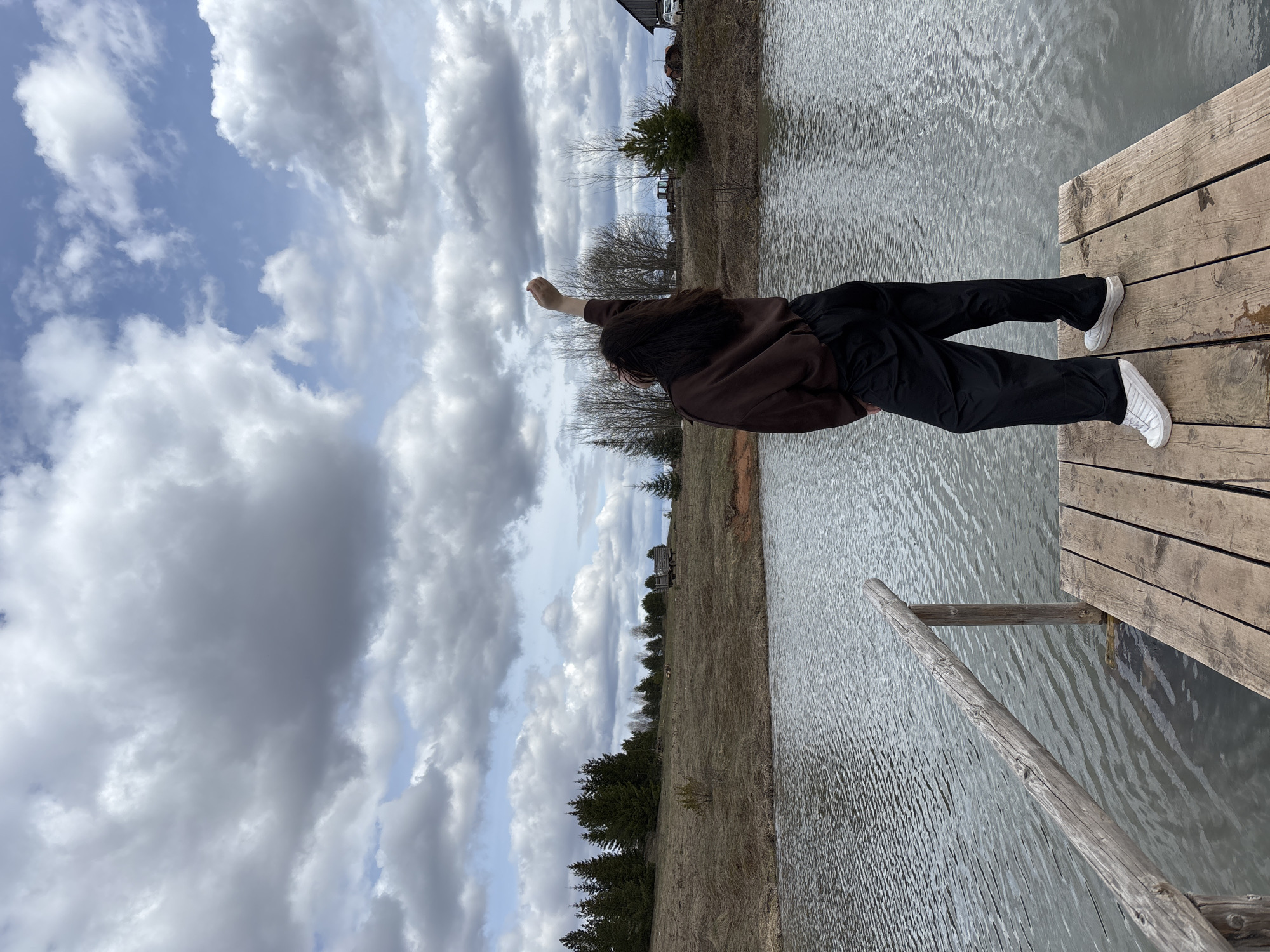 An outdoor medium shot captures a person standing on a wooden pier next to a body of water under a cloudy sky in what appears to be Petrograd, Russia. The image is oriented vertically with the person standing upright on the right side.
The main subject is an individual with long dark hair, wearing a dark brown long-sleeved top, black trousers, and white sneakers. They are positioned with their back mostly to the viewer, facing towards the left side of the frame, and are extending their right arm upwards and slightly outwards, with their hand open.
The person is standing on a weathered wooden pier or dock, constructed from light-colored planks. To the left of the pier is a calm body of water, likely a lake or large pond, which reflects the overcast sky with gentle ripples visible across its surface. A wooden railing or ladder extends from the pier into the water on the lower right.
The background consists of a natural landscape. The ground appears dry and grassy or covered with bare earth, suggesting an early spring or late autumn season. Several bare deciduous trees and some dark green evergreen trees (pines or firs) dot the shoreline. In the distance, on a slight elevation, a rustic-looking wooden building, possibly a log cabin or shed, is partially visible under the horizon line.
The sky is largely covered with an array of grey and white cumulus clouds, although patches of lighter blue sky are visible in some areas. The overall lighting suggests it is daytime, possibly late morning or afternoon. There is no visible text in the image.