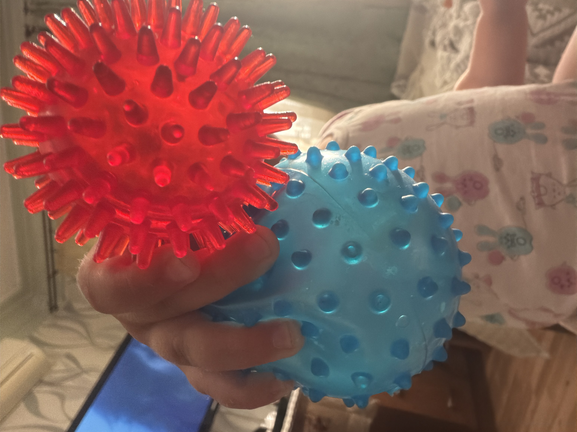 The image shows a close-up of a child's hand holding two spiky sensory balls, one red and one blue. The red ball is partially visible in the upper right, while the blue ball is in the foreground, held by the child's fingers. The child is wearing a light-colored shirt with a pattern of cartoon rabbits. In the background, a television screen is visible on the left, and a bed with patterned bedding is on the right. The scene appears to be indoors, possibly in a child's bedroom or living area. The lighting is somewhat dim, with light reflecting off the plastic balls. The image is taken from a low angle, looking up towards the child and the toys. It is not possible to determine the specific location within Komsomolskiy, Russia based on the visual information provided in the image.