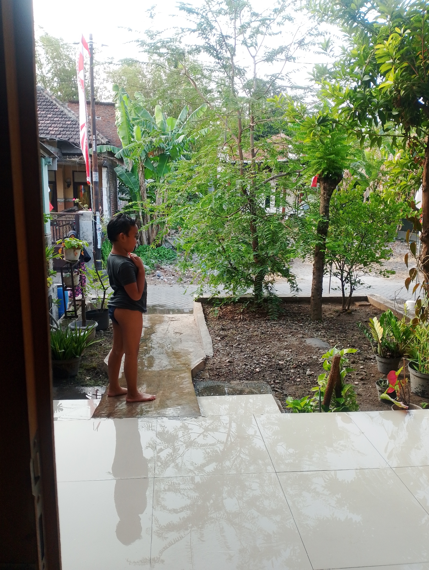 A young boy stands in profile on a wet concrete path outside a building, looking towards the right. He is wearing a dark t-shirt and what appear to be dark briefs. His hands are clasped in front of his chest. The ground is wet and reflective, showing a reflection of the boy and the trees.
The setting appears to be a residential area in Plumbungan, Indonesia. Behind the boy, a house with a tiled roof is visible, with a flagpole flying the Indonesian flag. The area is lush with greenery, including banana trees and other foliage. There are potted plants and gravel on the ground. The overall impression is of a warm, humid day, possibly after rain, given the wet surfaces. The boy's posture suggests a moment of contemplation or quiet observation. The scene is framed by the edge of a doorway or opening, with a wooden frame visible on the left.