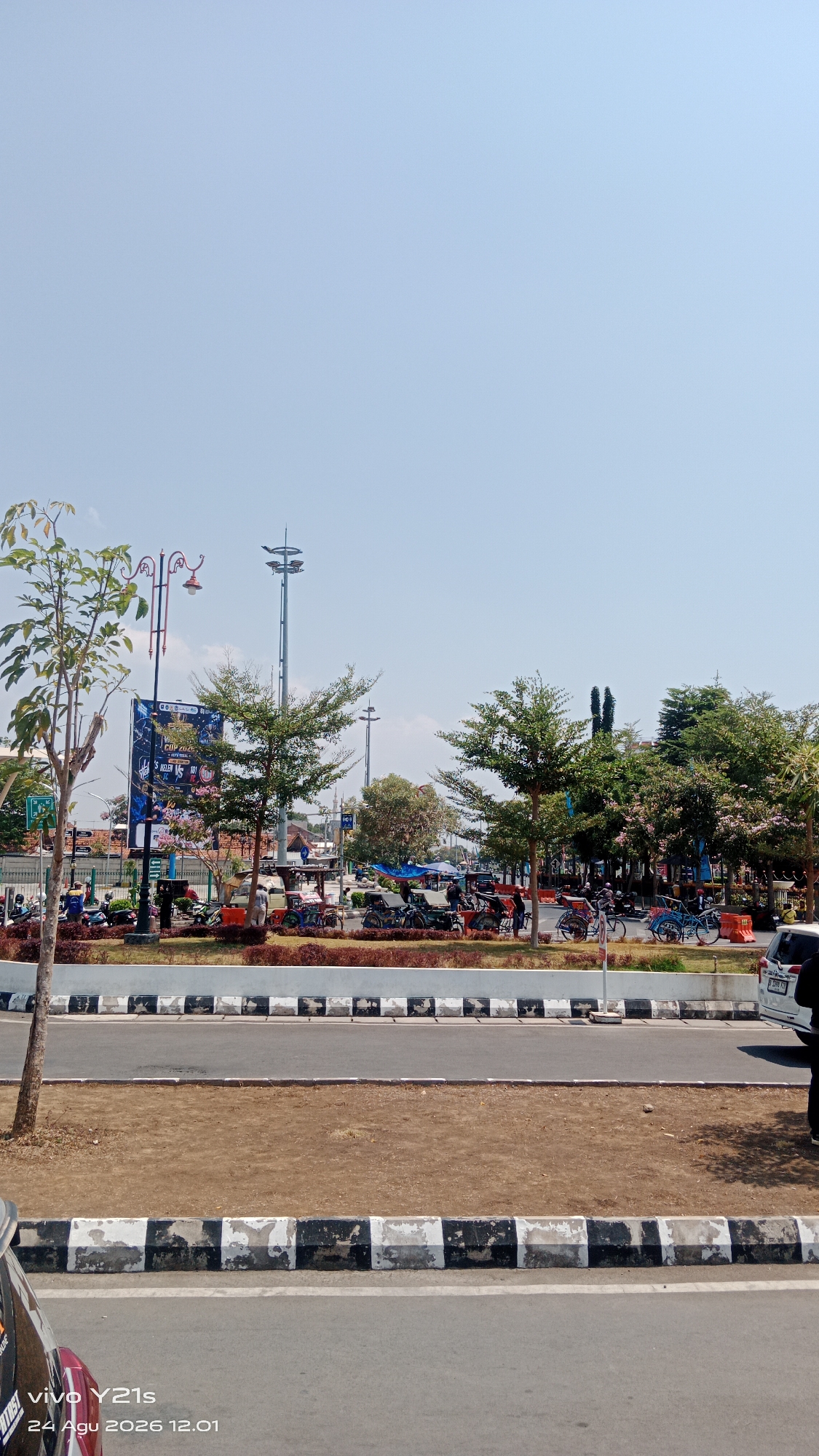 The image depicts a street scene in Tegal, Indonesia, on a clear day with a bright blue sky. In the foreground, a paved road is visible with a distinct black and white striped border. Beyond the road, a dirt area separates it from a landscaped median. This median features brown bushes and several small, green trees.
Further back, a raised sidewalk or pedestrian area is lined with more landscaping, including trees with sparse green foliage and flowering bushes with light pink blossoms. A large billboard is prominently displayed, featuring colorful graphics and text in Indonesian, advertising what appears to be a sporting event or competition. Several streetlights and decorative lamp posts are also visible.
The area behind the billboard is populated with a variety of vehicles, including motorcycles, bicycles, and what appear to be rickshaws or pedicabs, some with covered seating. Several people are seen interacting with these vehicles or walking in the area. In the distance, trees and buildings are visible, suggesting an urban environment. To the far right, the rear of a white vehicle is parked on the street. The bottom of the image shows a watermark indicating it was taken with a vivo Y21s on August 24, 2026, at 12:01.