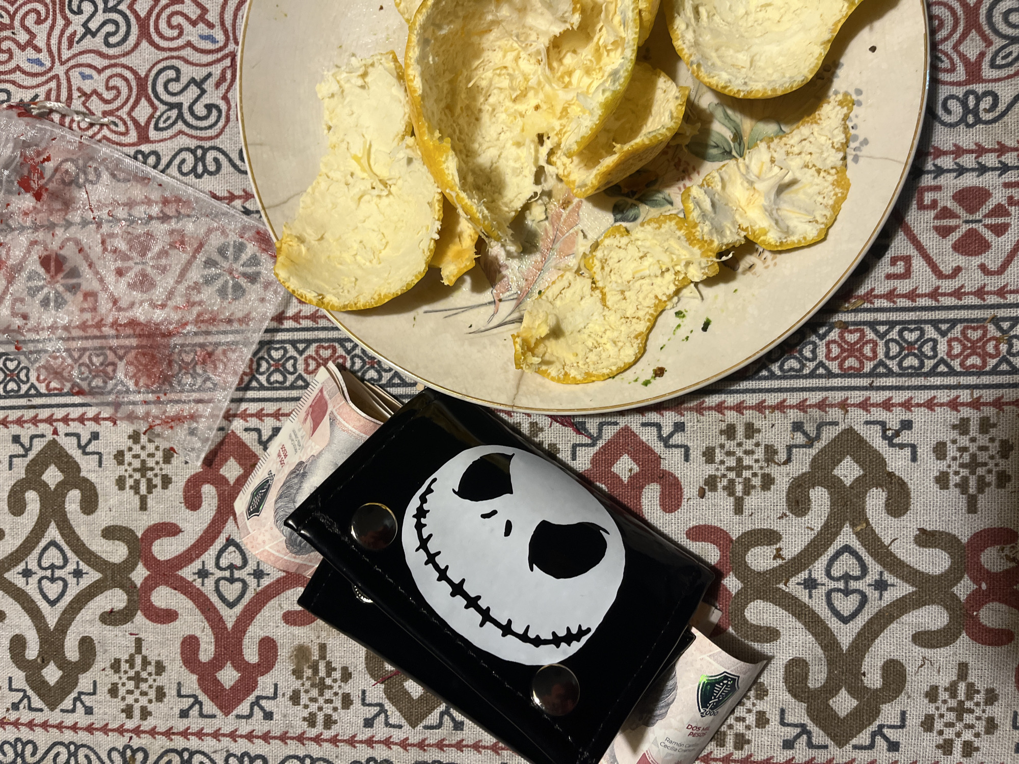 A close-up photograph shows a casual indoor scene. At the top of the image, there's a cream-colored plate with multiple segments of an orange peel. Below the plate, there's a wallet on top of money, showing the face of Jack Skellington. The background consists of a patterned tablecloth with geometric designs. The location is likely a private residence in Buenos Aires, Argentina, as indicated by the currency visible in the wallet. No people are visible in the scene, and the items suggest a moment of relaxation or routine domestic activity.
