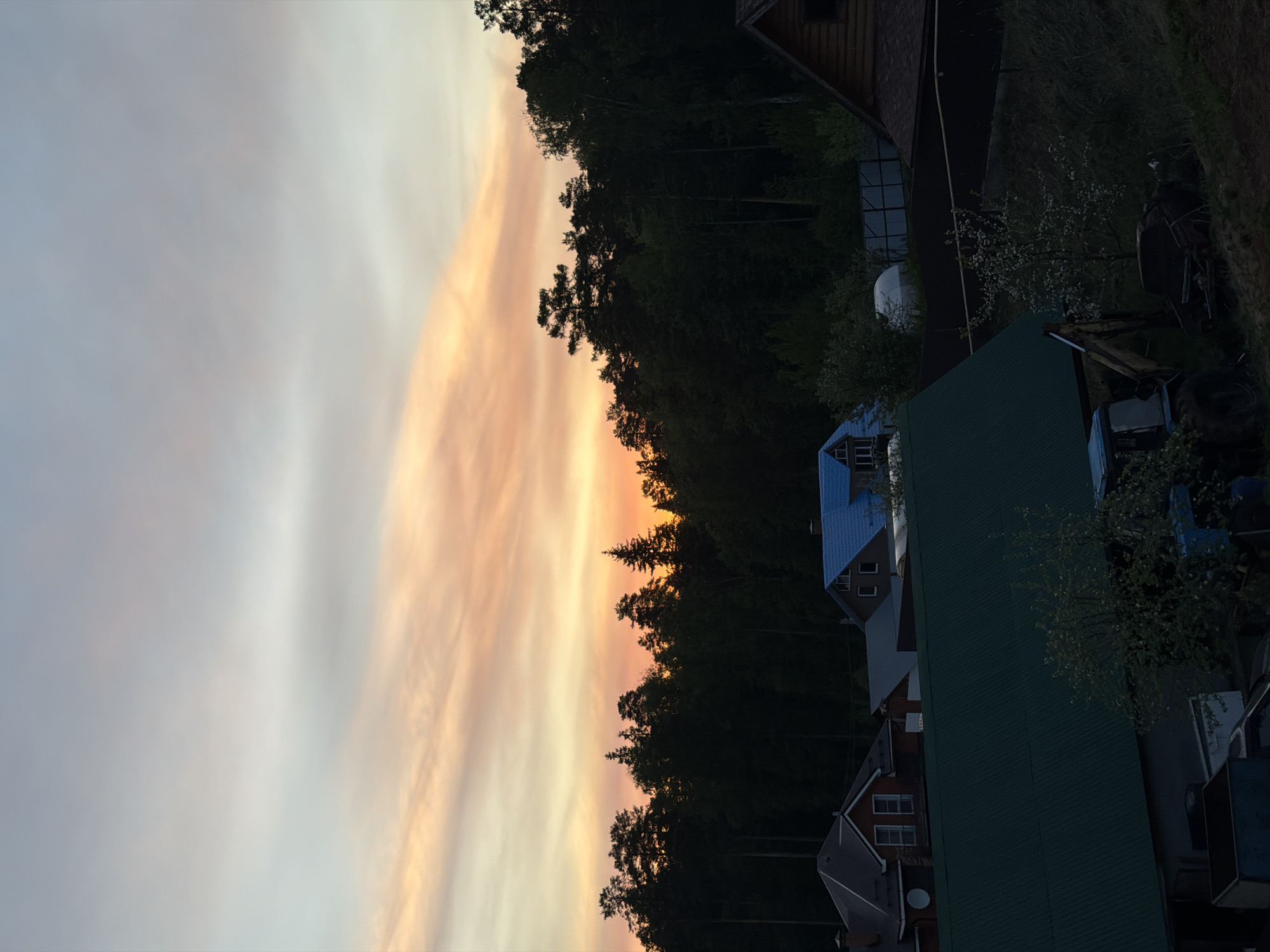 The image shows a sunset in a rural or suburban area, possibly Chaikovsky, Russia. The sky features a gradient of colors, from pale blues and grays to vibrant oranges and yellows, indicating either sunrise or sunset. The horizon is defined by a dense line of trees. To the right of the trees, houses and buildings with various roof colors (blue, green, brown, and grey) can be seen. There are also vehicles and other objects. The scene appears to be residential, situated near a wooded area. The lighting is muted, suggesting either early morning or late evening.