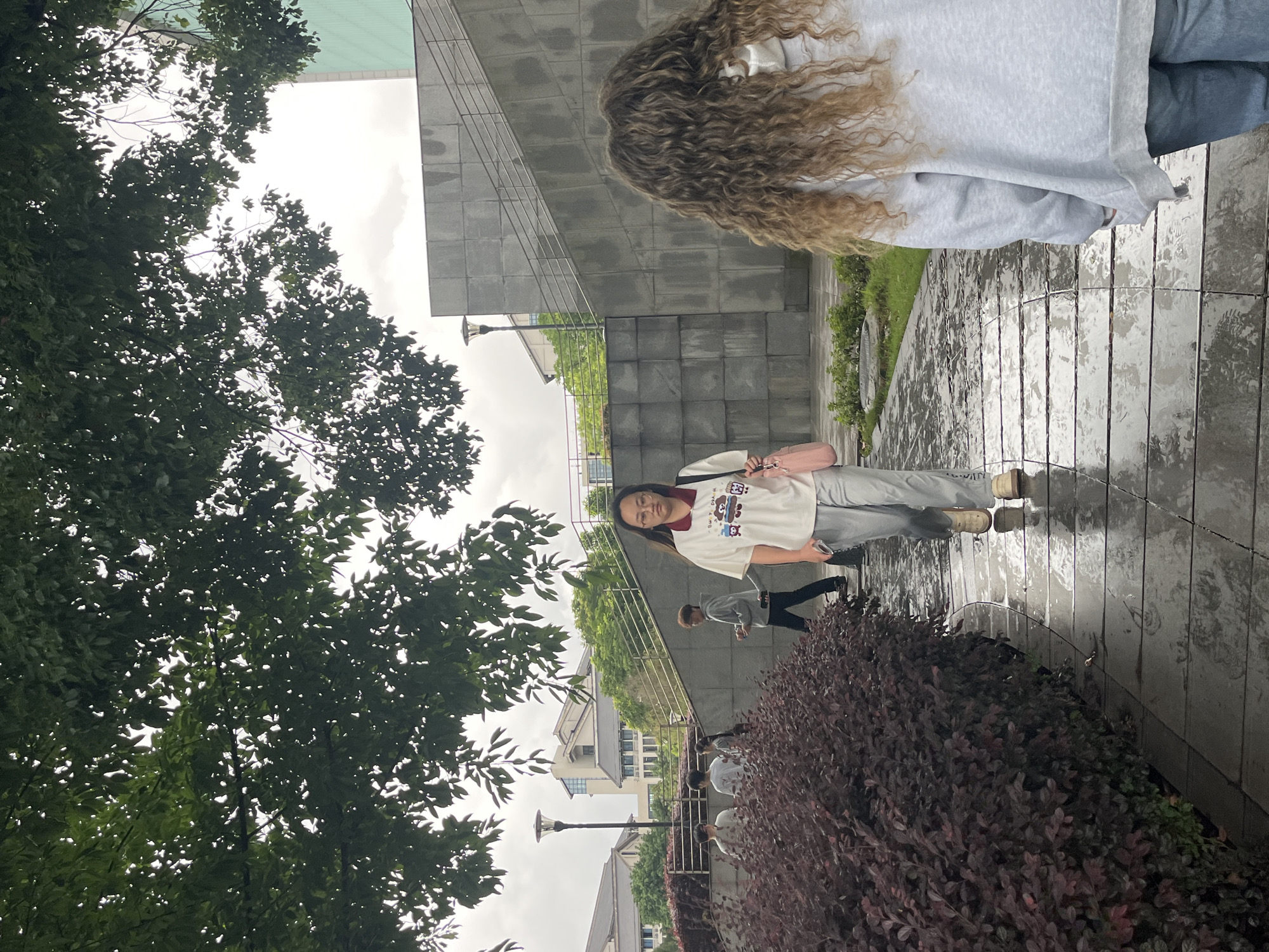 A group of people are walking on a wet, paved pathway in a park-like setting. The pathway is lined with dark red bushes on the left. In the background, there is a large, grey, angled concrete structure with stairs leading up it, and a light green building. Several people are visible, including a young woman in the foreground wearing glasses, a white t-shirt with graphics, and light grey pants, carrying a pink bag. Another person with long, curly blonde hair, wearing a grey hoodie and blue jeans, is walking away from the camera. Other individuals are seen in the mid-ground, walking in various directions. The scene appears to be outdoors on an overcast day, with wet surfaces suggesting recent rain. The architecture of the buildings in the background indicates an urban or campus environment. It is located in Jiangjin, China.