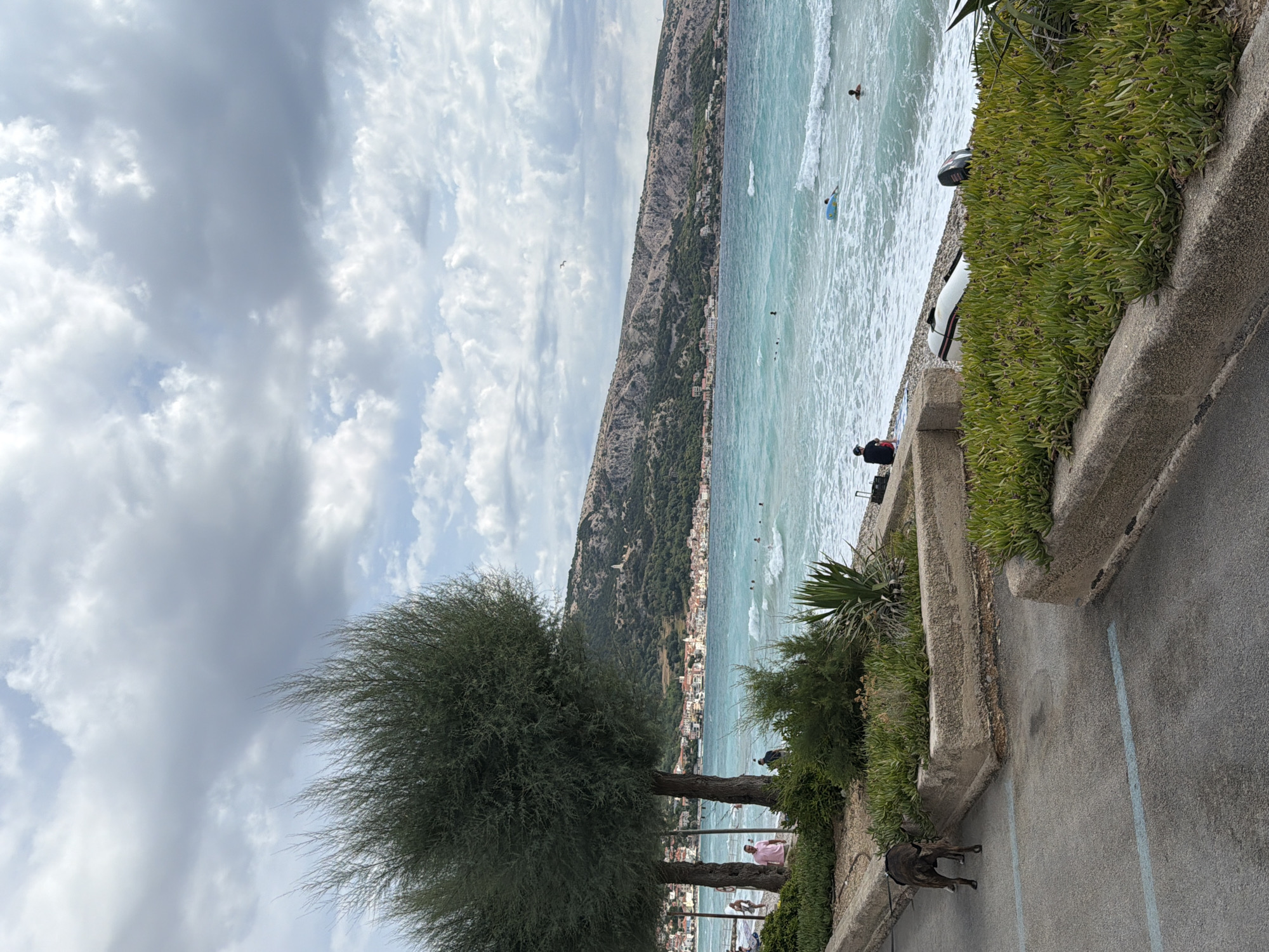 In Općina Baška, Croatia, a coastal scene unfolds with a clear sky and scattered clouds. The foreground features a paved area with a dog on a leash, facing away from the camera. Adjacent to the paved area is a raised planter bed filled with green succulents, bordered by a concrete structure. Beyond this, a pebble beach is visible, with several individuals swimming and enjoying the turquoise sea. In the background, a town with white buildings and red roofs nestles against a treed hillside, topped by a prominent structure. A small boat rests on the beach near the water's edge. The overall ambiance is one of a peaceful day at the seaside.