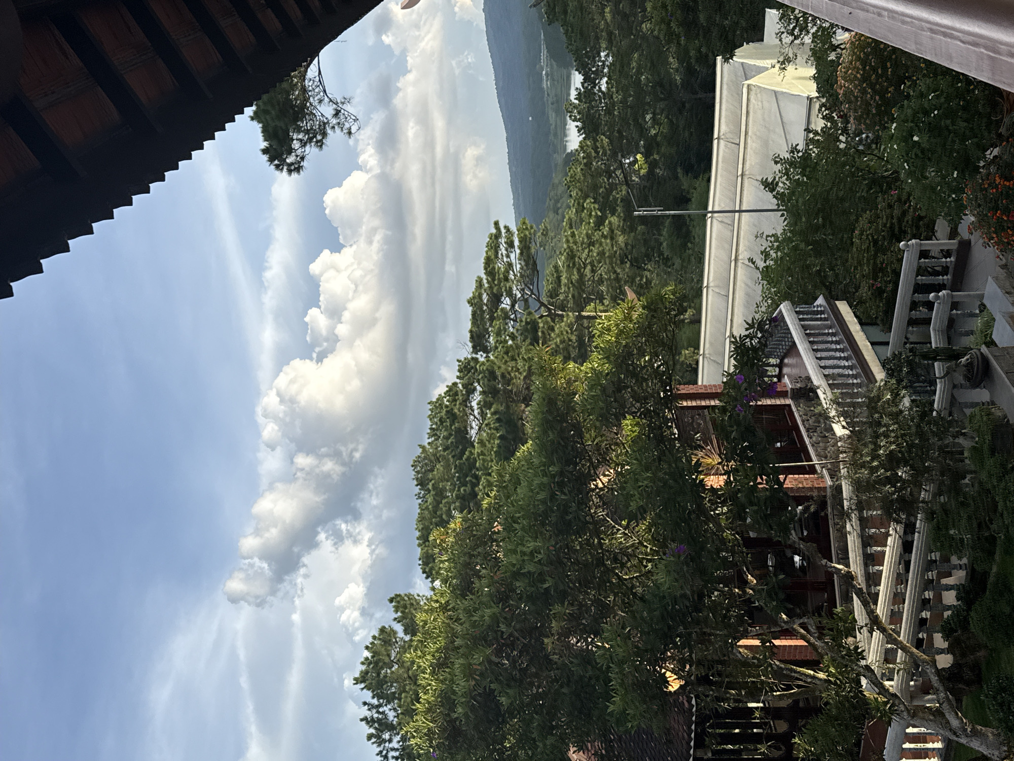 The image captures a view from a balcony in Da Lat, Vietnam, overlooking a lush landscape. In the foreground, a traditional Vietnamese house with a tiled roof and brick walls is visible, adorned with a white railing and surrounding greenery. Beyond the house, a series of greenhouses with white, translucent roofs are spread across the terrain. Further in the distance, a serene lake is nestled amongst rolling hills covered in dense pine forests, with a hazy sky dotted with cumulus clouds completing the scene. The overall impression is one of tranquility and natural beauty.