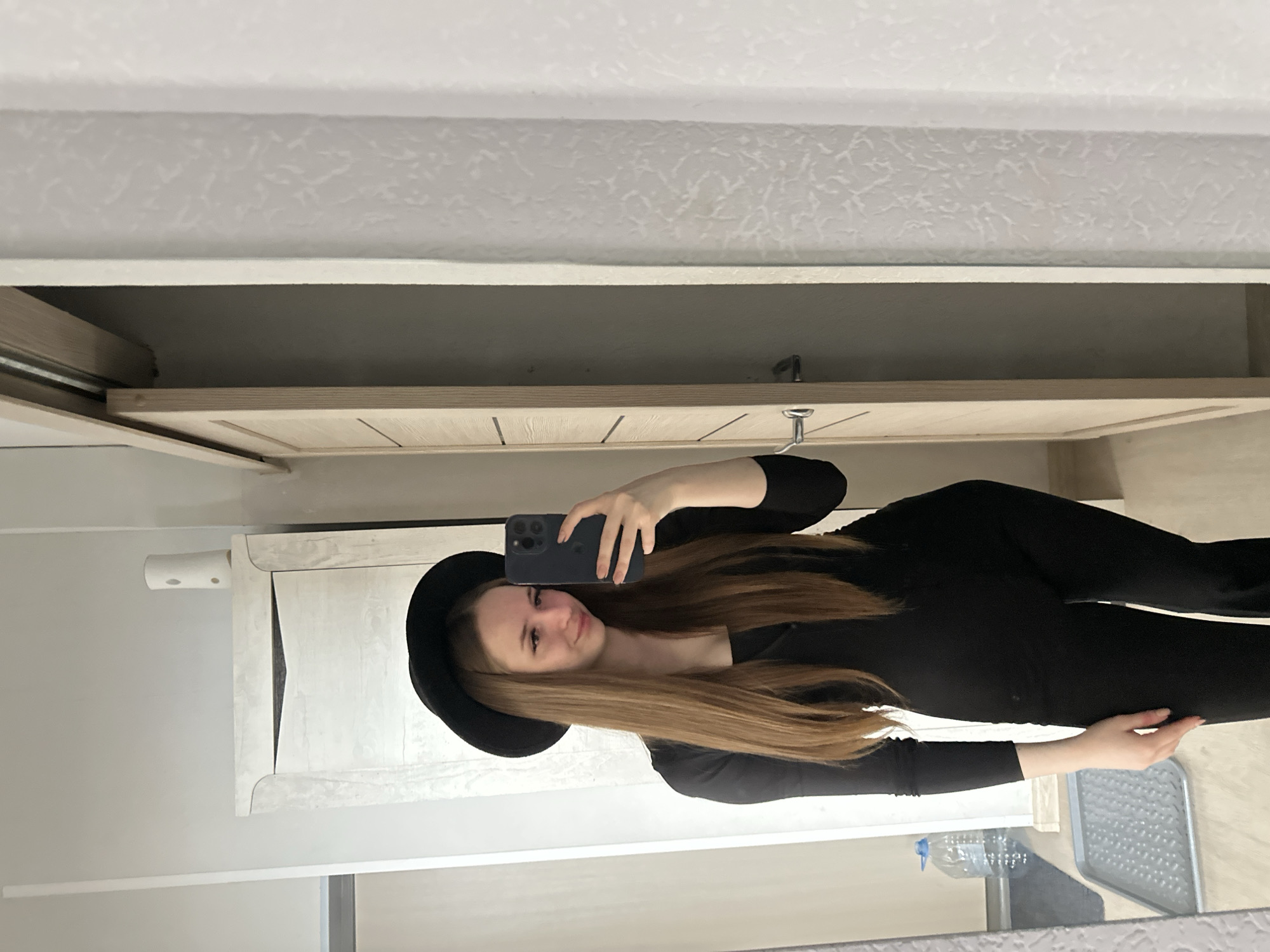A young person, likely female, is taking a selfie in what appears to be a bathroom or bedroom setting. She is wearing a black outfit and a wide-brimmed black hat, holding a smartphone up to capture the image. Her hair is long and light brown. The background includes a cabinet or wardrobe with a light-colored wood finish, white walls, and a patterned ceiling. There is also a glimpse of what looks like a metal stool and a water bottle. The exact location in the city of Unknown, Unknown is indeterminable from the image.