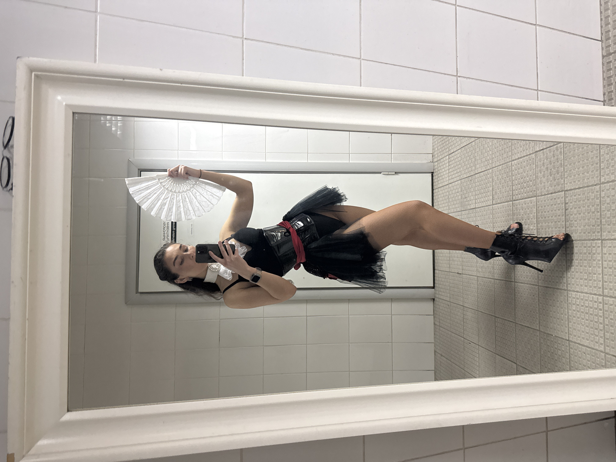 The image shows a full-length reflection of a young woman in a bathroom. She is wearing a black corset with a red sash, a short black tutu, black fishnet stockings, and black lace-up high-heeled ankle boots. She is holding a white fan in her right hand and her smartphone in her left hand, appearing to take a selfie. The bathroom has white tiled walls and a tiled floor. The mirror has a thick, ornate white frame. There is a door with a sign visible in the reflection. The scene is forensic and matter-of-fact. The location is specified as being in the city of Kazan, Russia, though this detail cannot be visually confirmed from the image itself.
