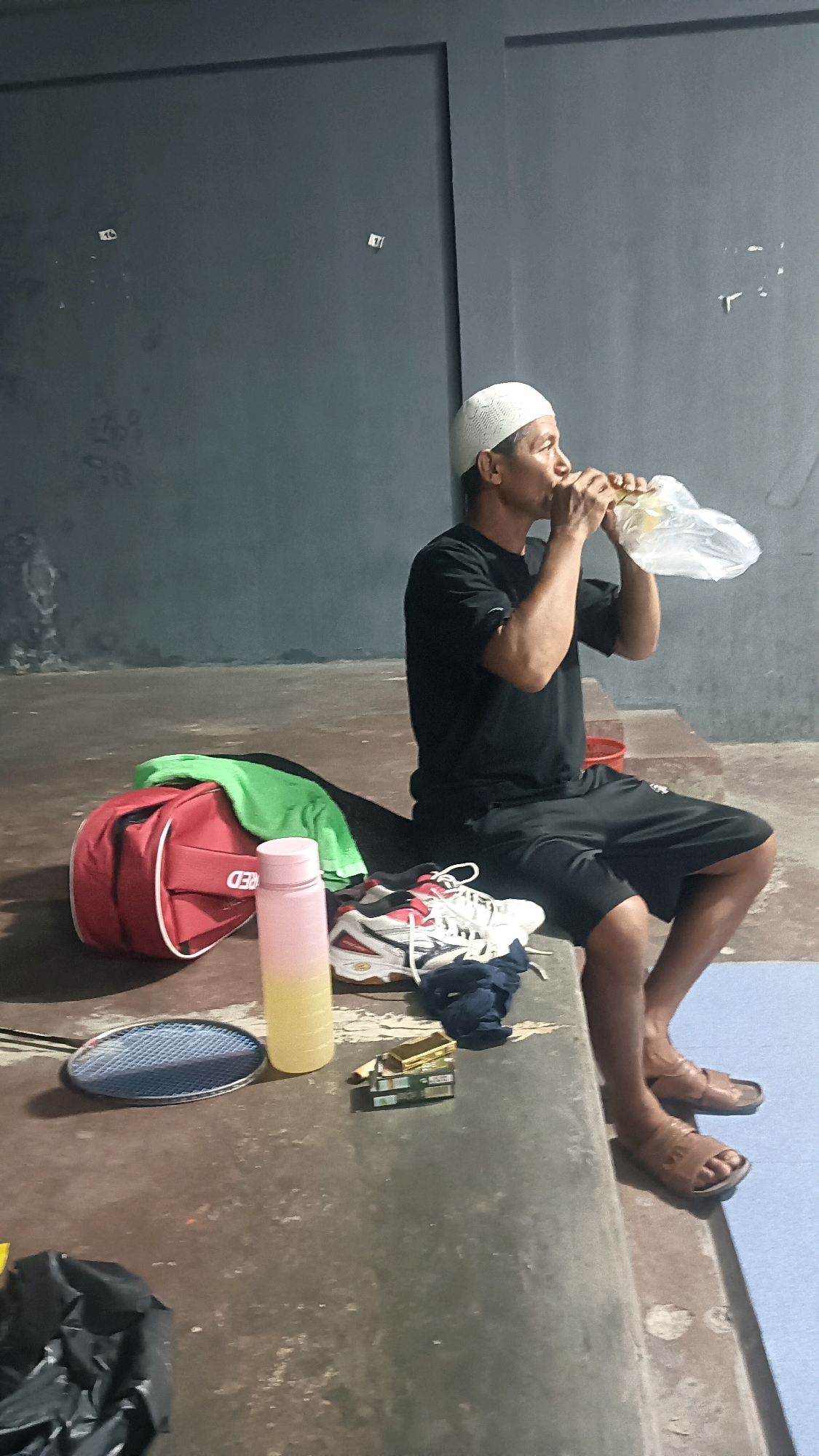 The image depicts a man wearing a white skullcap and a black t-shirt, seated on a concrete ledge and drinking from a clear plastic bag held to his mouth. He is in an indoor environment with a dark gray wall in the background, which has several small white rectangular labels or marks on it.
In front of the man, to his left, are several objects on the ledge. These include a red sports bag with a green cloth partially draped over it, a pink and yellow water bottle, a pair of white and red athletic shoes, and a dark blue piece of fabric. Further to the left and slightly lower is a blue badminton racket.
To the right of the athletic shoes, there is a small stack of what appear to be cigarette packs. The man is wearing black shorts and brown sandals. The floor in front of the ledge is a rough, textured surface, and to the far right, a section of light blue matting is visible. The scene appears to be a casual, possibly recreational, setting such as a badminton court or changing area. The location is identified as Tanggan, Indonesia.