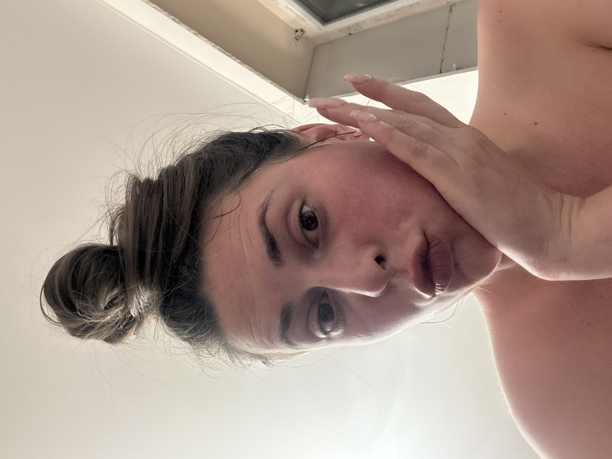 A woman with dark hair pulled into a messy bun poses for a selfie. Her right hand is on her cheek, and her lips are pursed. The background is a plain wall and a doorway. The lighting is bright and natural.