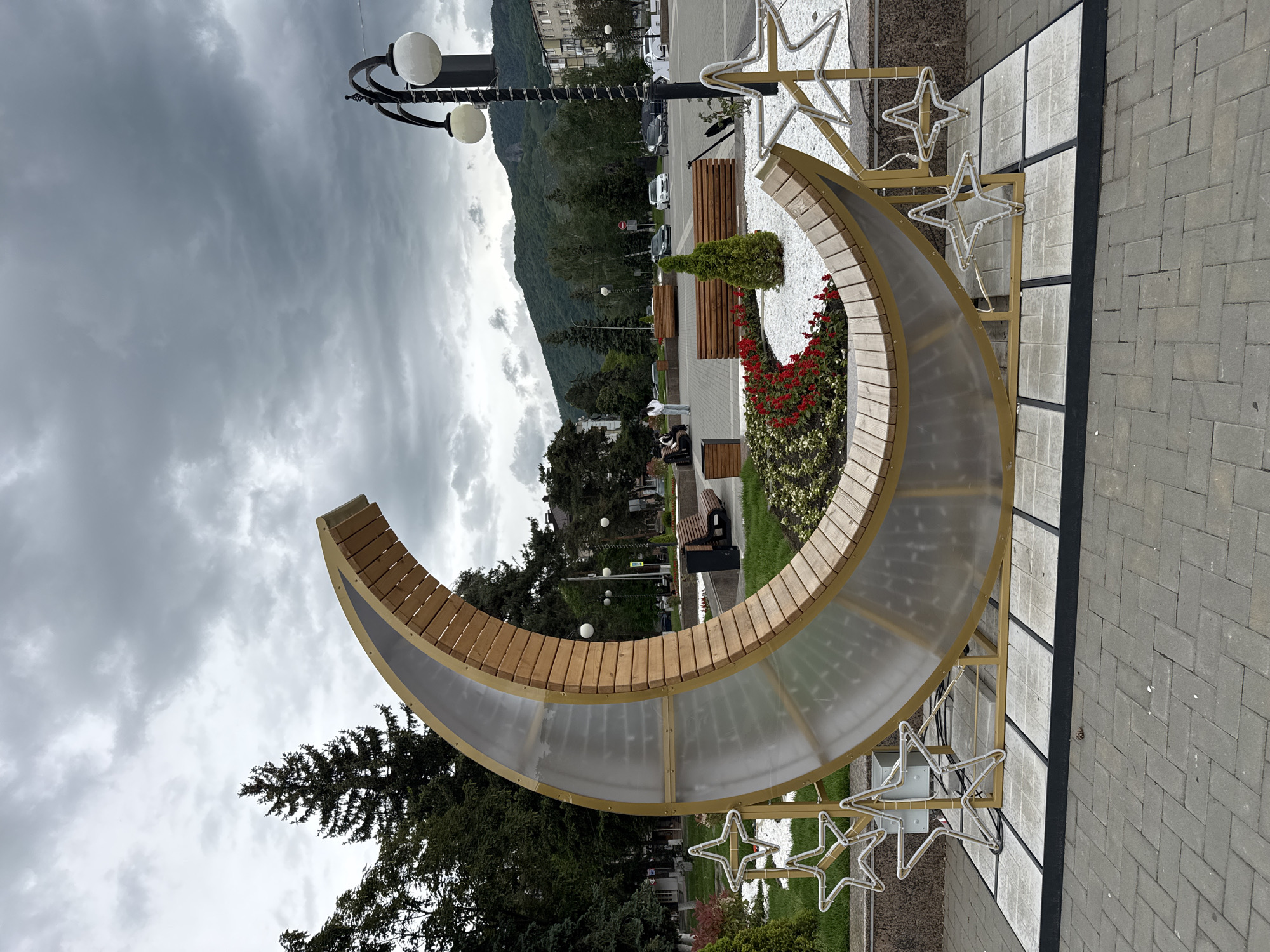 The image depicts a crescent-shaped sculpture in a public space in Karachayevsk, Russia. The sculpture consists of wooden slats forming the upper arc, supported by a translucent material below, all held together by a gold-colored frame. The sculpture is decorated with star-shaped lights. Inside the crescent shape, there is a display of red flowers and a small evergreen shrub, surrounded by white gravel.
In the background, there is a street with benches, greenery, and a lamppost. Further back, there are buildings and a mountain range under a cloudy sky. The ground is paved with grey brick. People are seen sitting on the benches.
