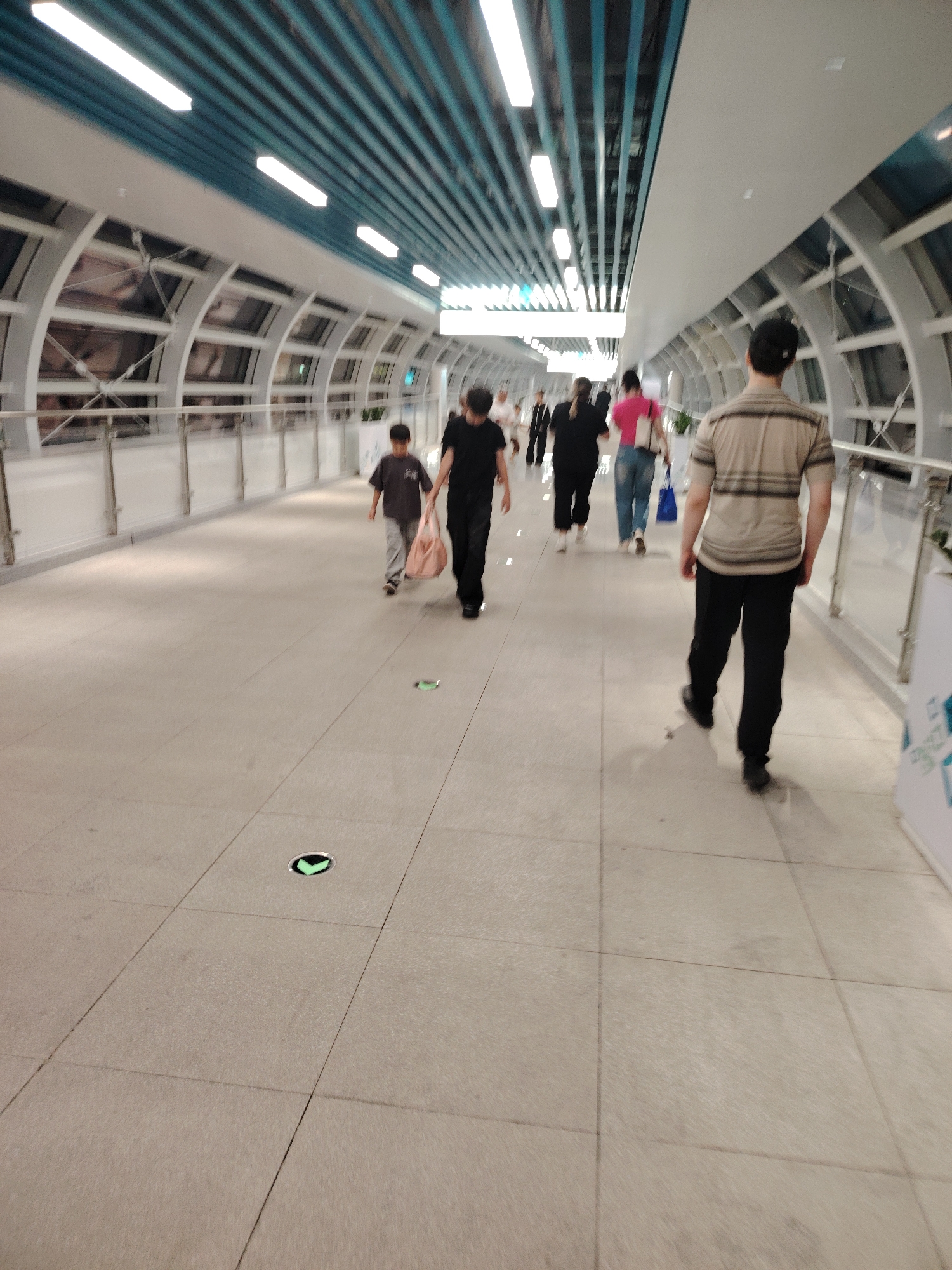 The image shows a group of people walking in a modern, indoor walkway. The walkway features a light-colored tiled floor, a white ceiling with blue striped accents, and large, arched windows that allow natural light to filter in. The architecture suggests a public space, possibly an airport, train station, or a large shopping complex.
Several individuals are visible, walking in the same direction. In the foreground, a man in a striped shirt and black pants is walking away from the camera. Further ahead, a child in a dark t-shirt and grey pants is walking, carrying a light-colored bag. Another adult, possibly accompanying the child, is walking behind them. Other individuals are also seen in the middle ground and background, some with blurred features due to motion.
The scene appears to be during the daytime, as indicated by the natural light. There are no visible weather cues. The overall atmosphere seems to be one of transit or casual movement. Two illuminated green arrow-shaped markers are visible on the floor, likely indicating directions or pathways. No discernible text is present in the image.