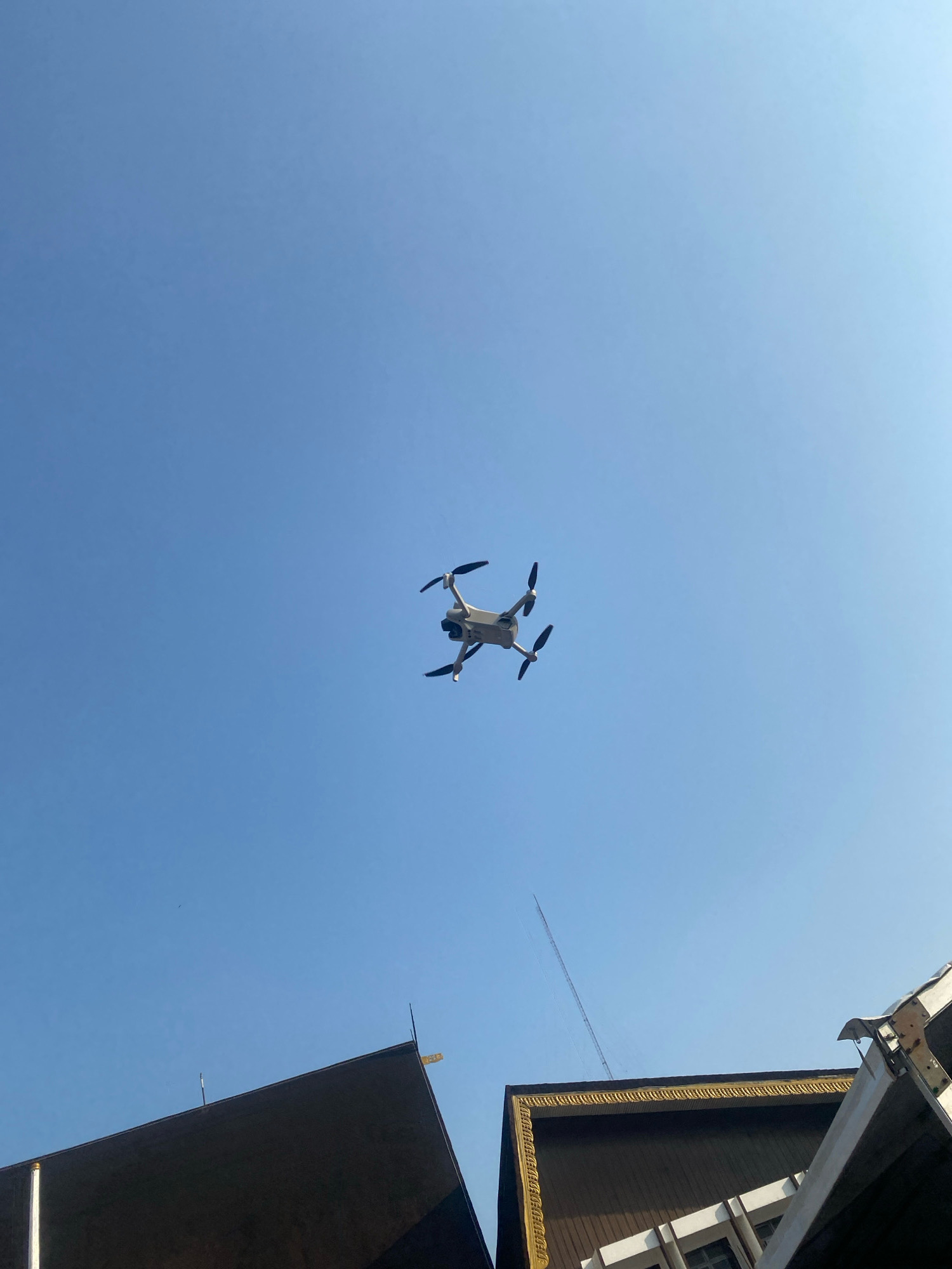 A white drone with four propellers is in flight against a clear blue sky. Below the drone, the tops of two buildings are visible. The building on the left is dark and angular, while the building on the right has a dark brown roof with a decorative gold trim and white accents. The image appears to be taken in Banjarmasin, Indonesia, given the architectural style of the buildings.