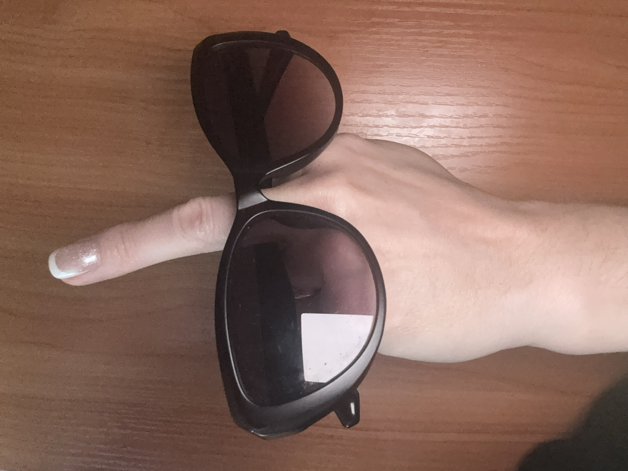 A top-down close-up shot shows a pair of dark-framed sunglasses resting on a light-skinned human hand. The hand is extended, with the index finger extended, lying underneath one side of the sunglasses. The sunglasses have dark lenses, one reflecting a light square. The background consists of a smooth, light-brown wooden surface. There is no indication of Tashkent, Uzbekistan.