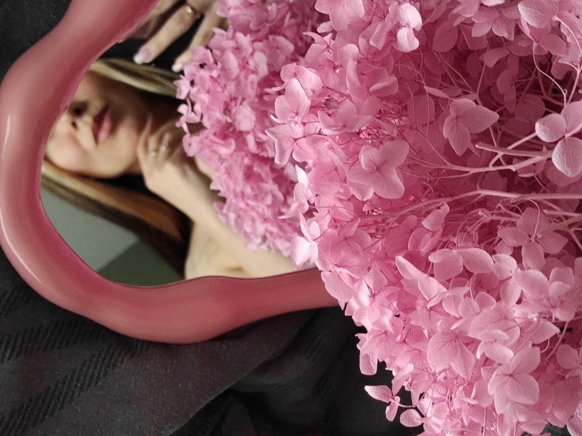 The image captures an artfully composed indoor scene, centered around a woman's reflection in a unique pink mirror, partially enveloped by a generous cluster of vibrant pink flowers. The mirror, featuring a glossy, irregular pink frame, occupies the left side of the frame, reflecting the softly blurred image of a woman. Her light-colored hair, delicate facial features, and hands, one of which displays rings, are visible. She appears to be gazing into the mirror with a gentle, contemplative expression.
Filling the right portion of the image and extending into the background is a dense arrangement of pink flowers, likely dried hydrangeas. These flowers are in sharp focus, highlighting their intricate, delicate petals and branches, and presenting various shades of pink. This sharp detail contrasts with the softer focus of the woman's reflection.
The mirror and the base of the flower arrangement rest upon a dark grey or black textured fabric, possibly a blanket or sweater, visible in the foreground and partially in the background. The overall aesthetic is one of softness, femininity, and introspection, achieved through the interplay of colors, textures, and depth of field. There are no explicit indicators for the time of day or specific location cues within the image, nor is any text visible.