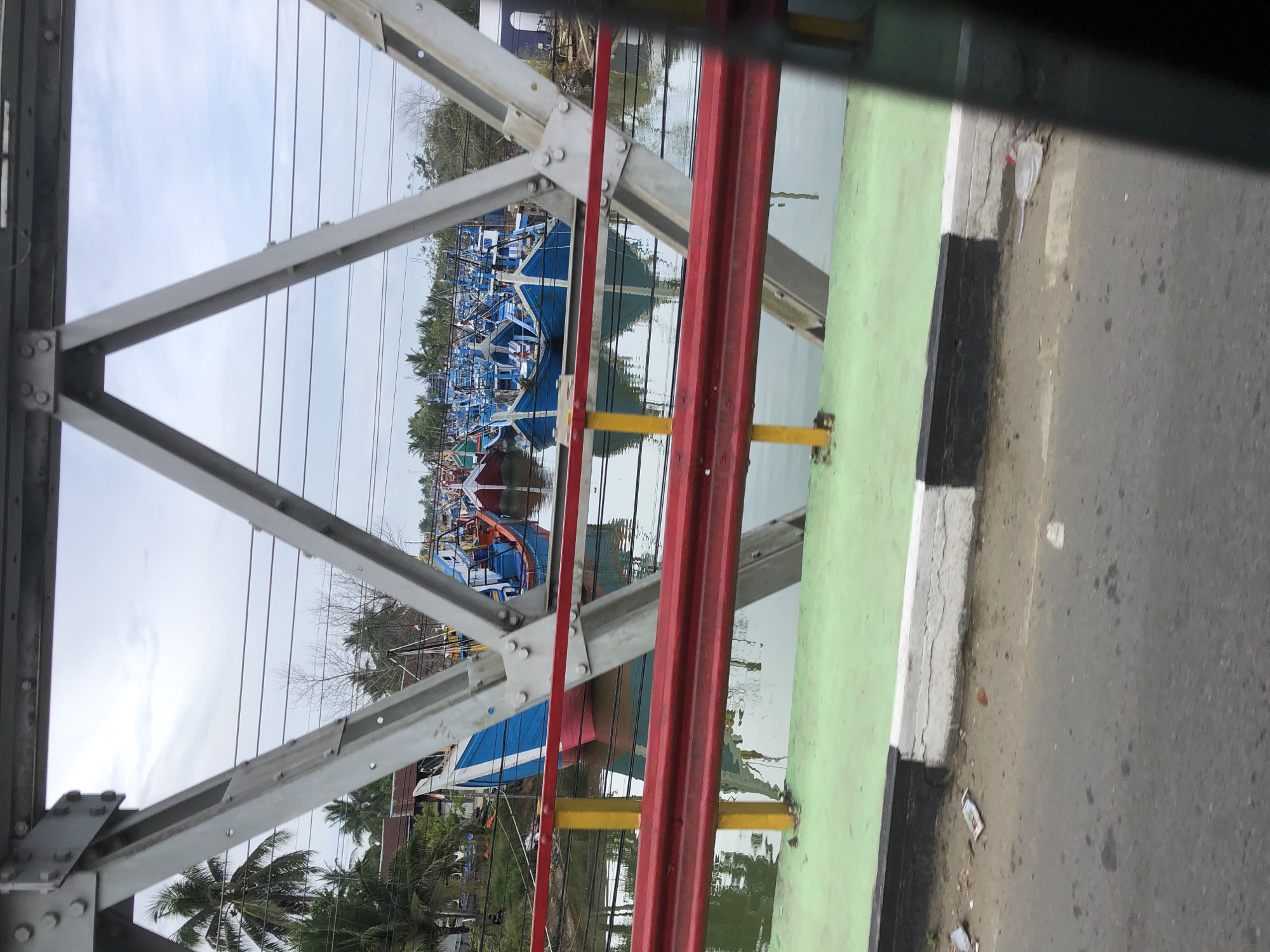 From a roadside vantage point on a bridge, the image shows a view of boats on the water in Idi Rayeuk, Indonesia. The bridge is constructed of gray metal beams and red guardrails with yellow supports. The curb adjacent to the road is painted with alternating black and white sections. On the road surface, there is some discarded paper trash. Through the bridge structure, numerous blue and red boats are visible on the water. Palm trees and other vegetation line the water's edge. The sky is overcast.