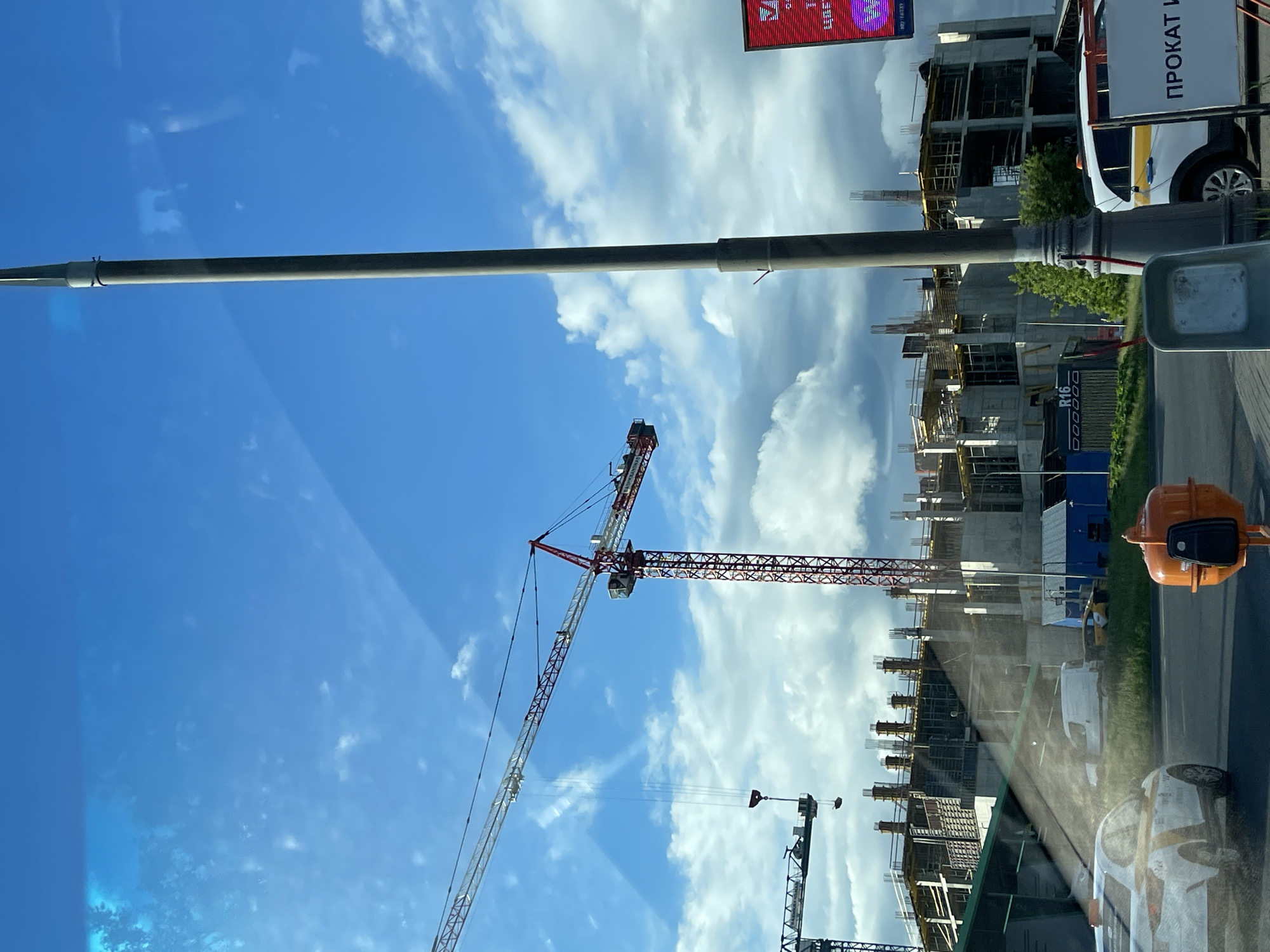The image shows a construction site in Bittsa, Russia. Several high-rise buildings are under construction, with construction cranes towering over them. The sky is blue with scattered clouds. Street furniture is present in the foreground, including traffic signals and a lamppost. Vehicles, including a van with Russian writing on the side, are parked or driving on the road adjacent to the construction site.