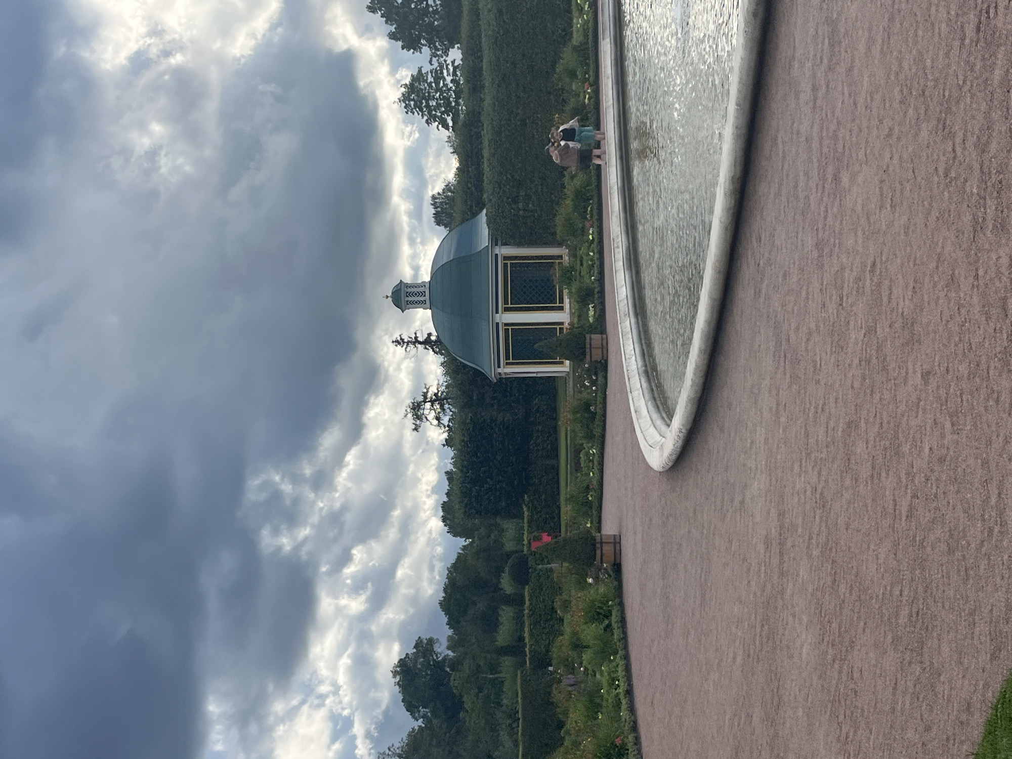 In Peterhof, Russia, a circular fountain with a stone rim is situated on a pathway made of reddish gravel. To the left of the fountain, a curved path leads further into a manicured garden featuring hedges, flower beds, and potted trees. In the background, a small, ornate green and white gazebo stands amidst dense green hedges. Two individuals, a man and a woman, are standing by the edge of the fountain, looking out at the water. The sky above is overcast with dark, dramatic clouds.