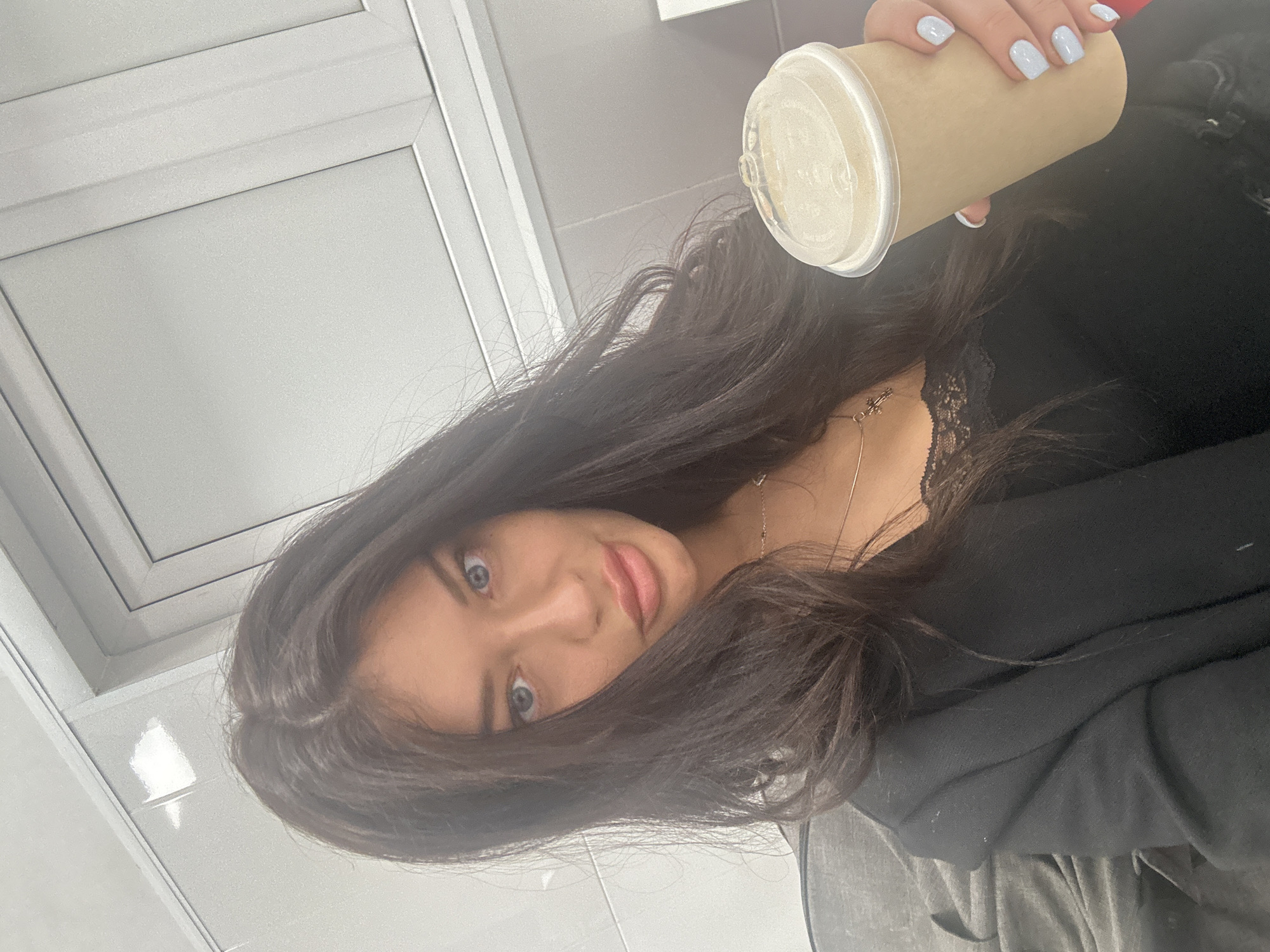A young woman is featured in a close-up selfie, taken from a low angle and rotated 90 degrees clockwise. She appears to be looking directly into the camera with a subtle, pleasant expression.
The main subject is a woman with long, dark brown hair that cascades around her face and shoulders. She has striking blue eyes and light pink lips. She is wearing a black top, possibly a sweater or jacket, with a visible touch of black lace trim at the neckline. A delicate silver-toned necklace with a small pendant is visible around her neck.
In her right hand, she holds a light brown, disposable cup, likely made of paper or cardboard, with a clear plastic lid. Her fingernails are painted a soft, light blue color. Faint, unreadable text or a logo is present on the clear plastic lid of the cup.
The background is a minimalist indoor setting, characterized by light gray or white rectangular panels that suggest a modern, clean environment, possibly an office, retail store, or contemporary interior. The panels are geometrically arranged, forming lines and squares. The lighting is bright and even, indicating an indoor setting with artificial light.
The activity taking place is a self-portrait. The woman's direct gaze and slight smile convey a sense of confidence or contentment. No specific time of day or weather can be determined from the indoor setting. There is no clearly legible text on any visible objects, beyond the faint markings on the cup lid.