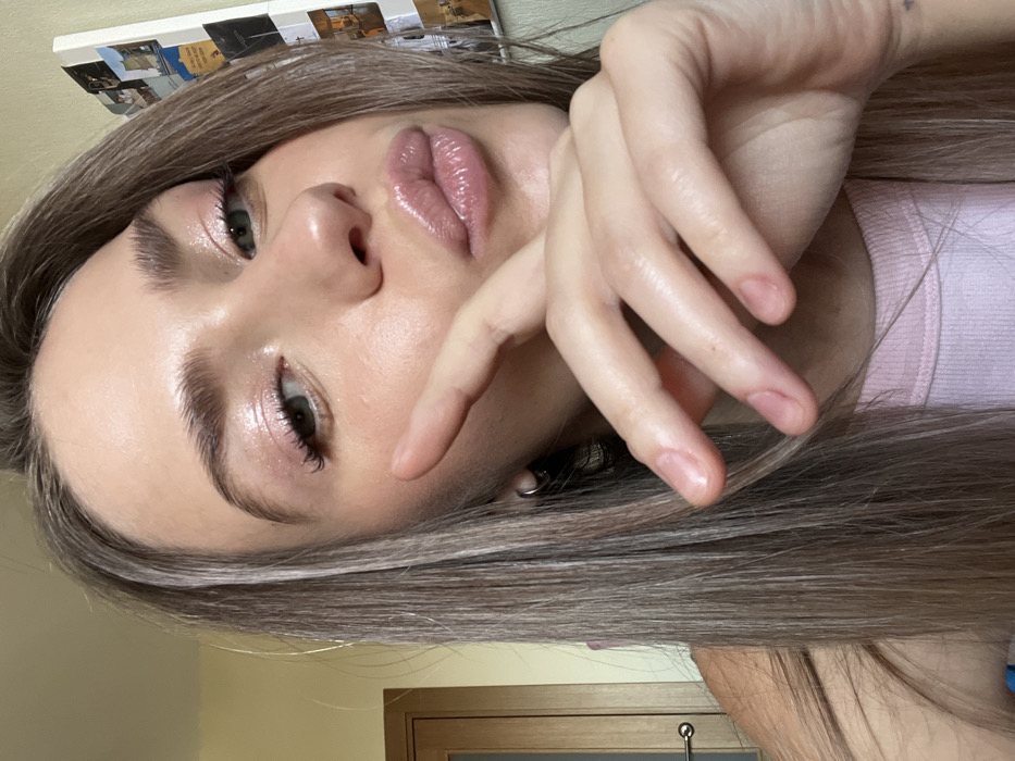 A close-up selfie captures a young adult female from the chest up in an indoor environment, presumed to be in Saint Petersburg, Russia. The individual has light-toned skin and long, straight light brown hair with visible blonde highlights. She wears a light pink ribbed tank top. Her left earlobe displays a small, silver-toned hoop earring. Her face features makeup, including shimmery pink eyeshadow, defined eyebrows, and glossy pink lips, which are pursed in a "kiss" pose. Her right hand is partially visible, raised with fingers gently curled, and the index finger extends forward, resting near her mouth. Her fingernails are natural and unpolished.
The background elements include a section of a light-colored wooden door with a horizontal metallic handle positioned on the left side of the frame. On the right, a white vertical surface, possibly a refrigerator or magnetic board, is visible, adorned with several small, rectangular items that appear to be photographs or notes. The specific location within Saint Petersburg cannot be ascertained from the visual details provided in the image.
