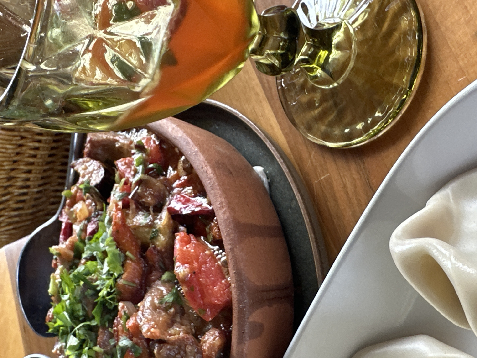 A table in Marneuli, Georgia, holds a feast: a clay pot filled with a stew of tomatoes, meat (possibly lamb or beef), and herbs; a plate of dumplings; and an amber-colored wine glass. A woven basket is slightly visible at the far left edge. The scene is indoors, likely a restaurant.
