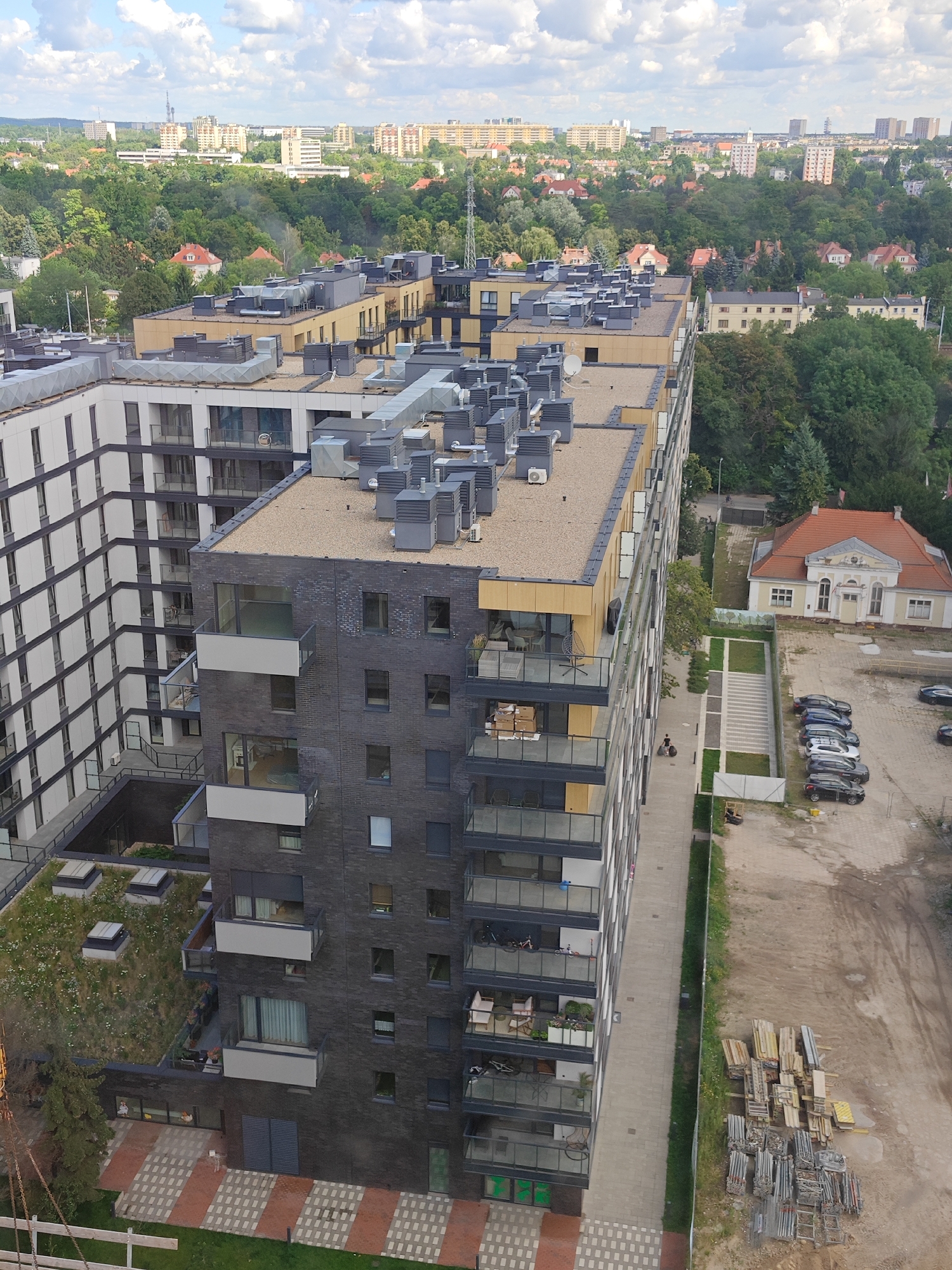 An aerial, high-angle, eye-level shot captures a modern, multi-story apartment building in Poznań, Poland, on a cloudy day. The building, constructed with dark brick and white accents, features numerous balconies with glass railings and gray air conditioning units on its flat roof. Adjacent to it, another taller building with a white facade and dark trim is visible, also with multiple balconies. The scene is surrounded by lush green trees and a distant cityscape with more buildings. In the foreground, a patch of green grass and a decorative brick pathway are visible. To the right of the apartment building, a small, pale yellow house with a red roof stands, and next to it, a gravel lot is filled with parked cars and stacks of construction materials. A person is walking along a paved path next to the apartment building. The sky is partly cloudy with hints of blue.