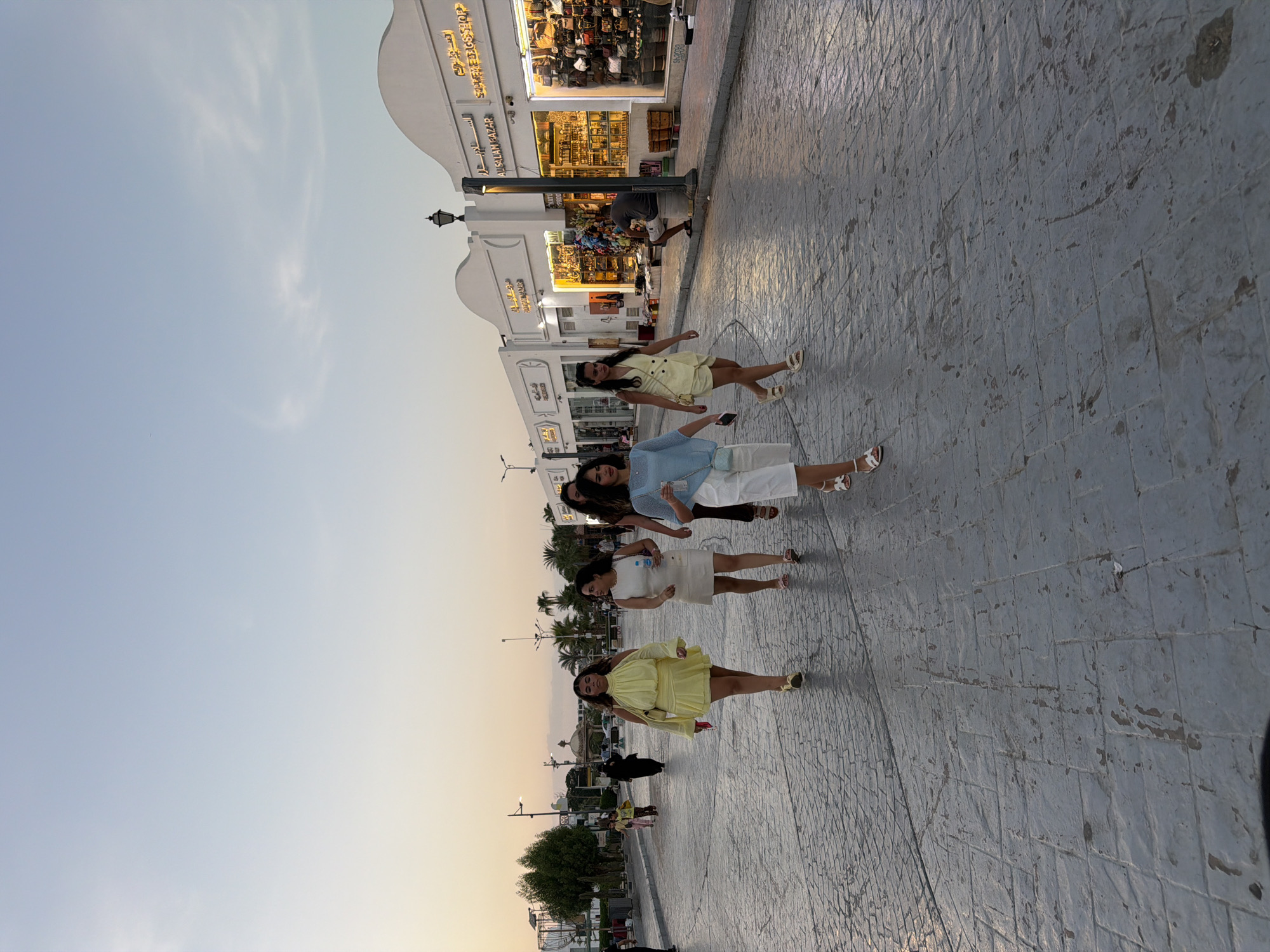 The image shows a street scene in Sharm El Sheikh, Egypt, likely during the early evening. A group of four young women are walking towards the viewer on a paved cobblestone street. The woman in the foreground on the left is wearing a bright yellow dress with flared sleeves and a matching mini skirt. The woman next to her is wearing a white dress and carrying a water bottle. To their right, a woman in a light blue top and white shorts is walking, and another woman in a yellow suit is walking slightly behind her.
In the background, there are shops with arched entrances displaying merchandise. One shop sign reads "AL SALAM BAZAR" and another says "SUPER BAGS SHOP". People are visible inside the shops and walking along the sidewalk. A man is seated on a low structure in front of one of the shops. Palm trees and other greenery line the street, and there are streetlights and architectural elements suggesting a tourist-oriented area. The sky is a pale blue, transitioning to orange at the horizon, indicating dusk.