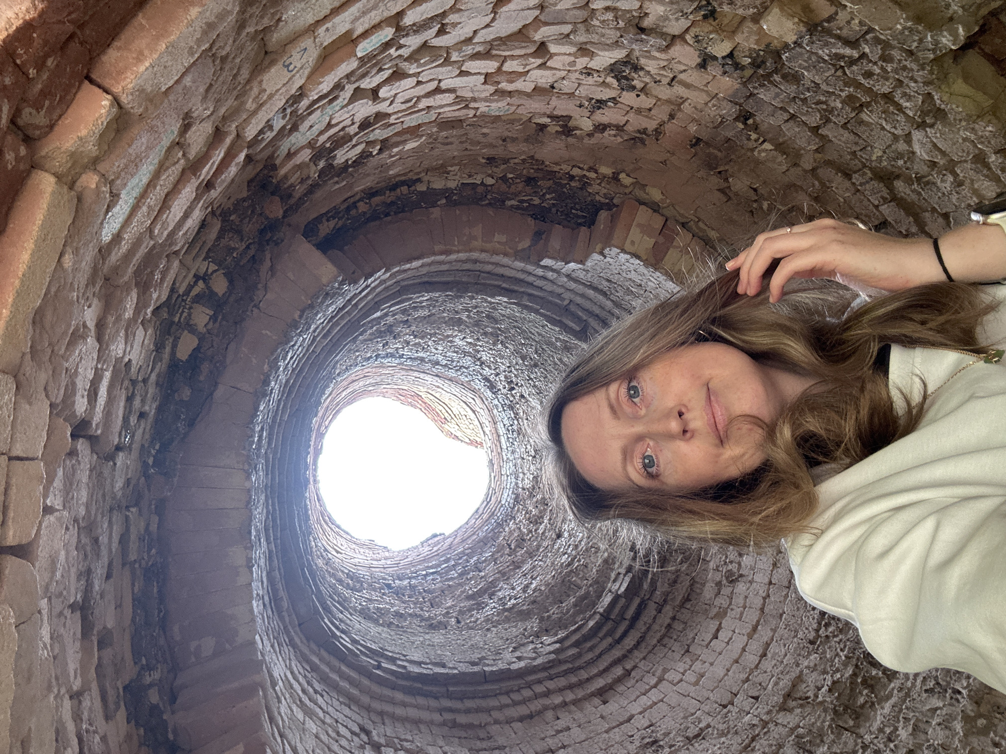 This media file is a selfie taken by a woman inside a brick kiln in Mezhevoy, Russia. The woman is positioned in the upper right of the frame, with her face turned towards the camera. She has light brown hair and blue eyes, and is wearing a light-colored hoodie. Her left hand is resting near her hair. The kiln is circular and constructed of rough, aged bricks, with the interior curving inwards and upwards to an opening at the top, through which daylight streams in. The bricks appear weathered and in some areas, discolored. There is some faint blue-green coloring on some of the bricks, and what appears to be faint numbering or markings on a few bricks in the upper left portion of the image. The perspective is from below, looking up through the kiln towards the opening. The image suggests an indoor setting, likely during the daytime given the bright light from above. The woman's expression is neutral, with a slight smile. The scene evokes a sense of exploration and curiosity.