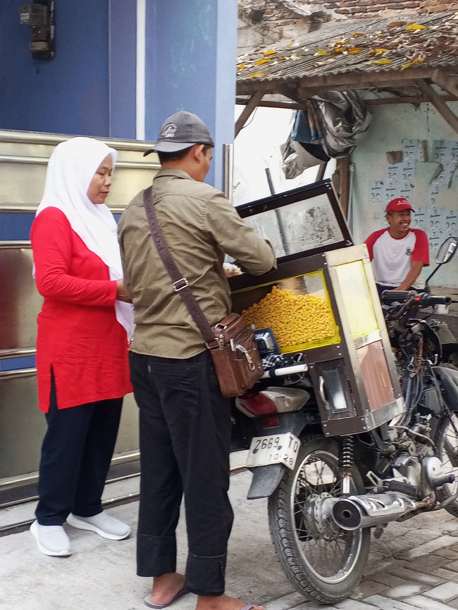 In the city of Ciruas, Indonesia, a street scene unfolds with a woman and two men interacting near a motorcycle food cart. The woman, dressed in a vibrant red long-sleeved shirt and a white hijab, stands on the left, wearing white sneakers. To her right, a man in a khaki long-sleeved shirt, dark pants, and a grey baseball cap, faces away from the camera, carrying a brown shoulder bag. He appears to be a vendor, serving food from a yellow and brown cart mounted on a motorcycle. The cart contains a large pile of yellow snacks, possibly corn puffs. To the right of the food cart, a second man wearing a red and white t-shirt and a red baseball cap is seated on the motorcycle, smiling and looking towards the cart. The background reveals a building with blue walls and a corrugated metal roof structure over the food cart, suggesting a roadside establishment. The motorcycle, a key element of the scene, has a license plate reading "2669. TD 10. 28". The overall impression is a snapshot of daily commerce and human interaction in an Indonesian urban setting.
