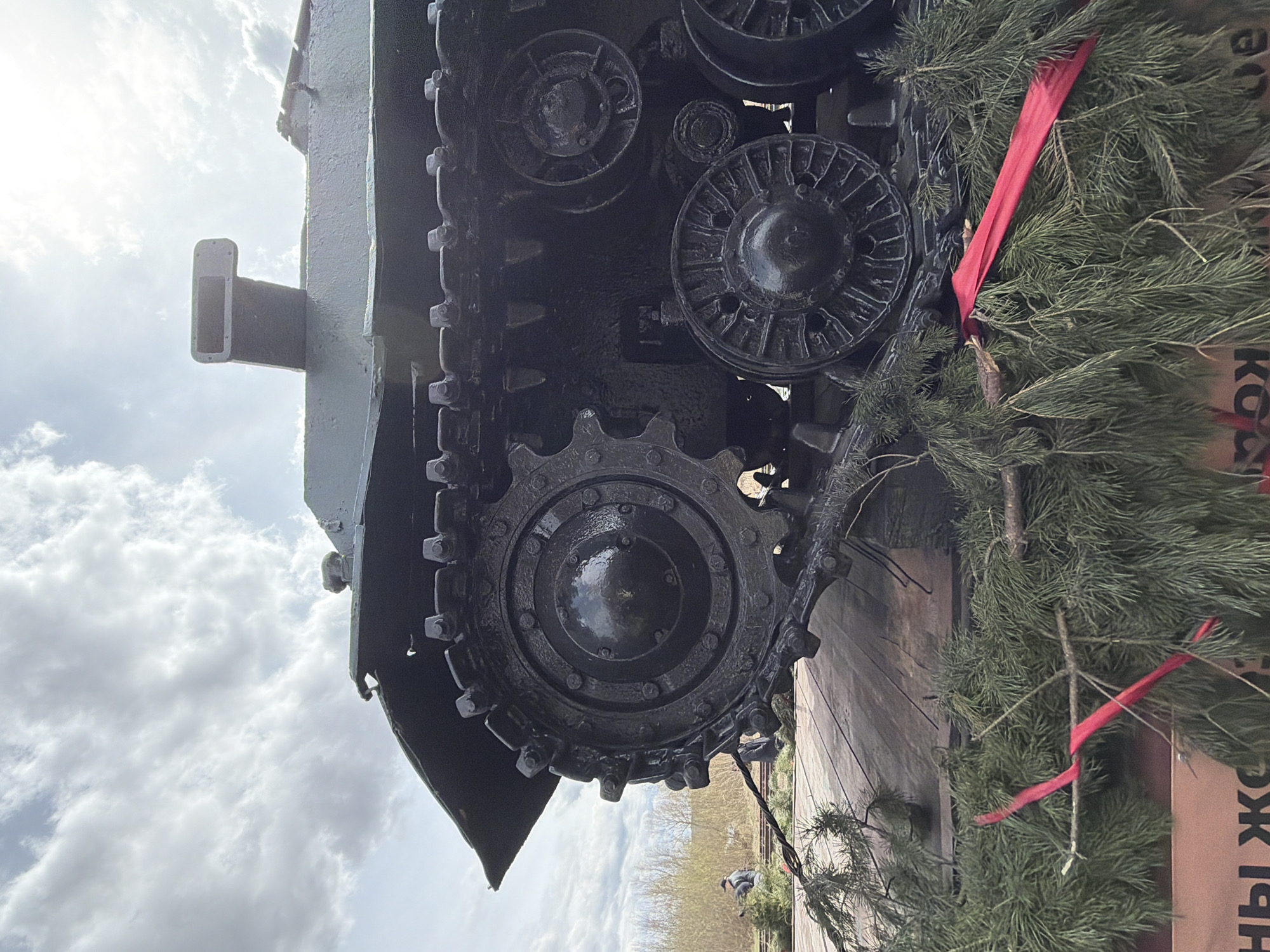 The image presents a close-up view of a military tank, oriented vertically on its left side. The tank's track system dominates the central and right portions of the frame, featuring a large, dark sprocket wheel with intricate details, along with several smaller road wheels and heavy track links. The metal components are painted a dark, possibly black or very dark green, color, showing some texture and worn highlights.
A prominent rectangular structure, resembling a snorkel or exhaust, extends horizontally from the upper part of the tank's hull, visible against a bright, cloudy sky that occupies the left side of the image. The sky is largely overcast but luminous.
On the right side of the track system and extending towards the bottom right, numerous fresh green pine branches with needles are arranged, suggesting a decorative or commemorative display. A vibrant red ribbon is tied around some of these branches. Below the track system and obscured by the foliage, a section of a light brown surface with visible Cyrillic text can be seen, partially revealing the letters "ЖК" and "ЫН", likely part of a Russian word or sign. The ground below the tank appears to be a wooden platform. The scene takes place during the daytime under bright, diffused lighting, suggesting an outdoor exhibit or monument in Votkinsk, Russia. No people are visible.