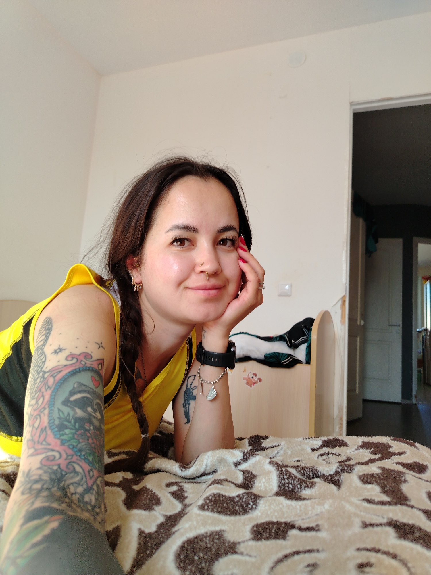 The image shows a young woman lying on a bed with her left arm extended towards the camera. She is wearing a yellow and black tank top and has several tattoos on her left arm. Her hair is dark brown and braided. She is resting her head on her right hand and looking at the camera with a slight smile. The room appears to be a bedroom, with a bed covered in a patterned blanket. In the background, there is a door and part of a room with dark flooring. A white bedside table with some items on it is visible to the right of the woman. The location is identified as Noviy, Russia.