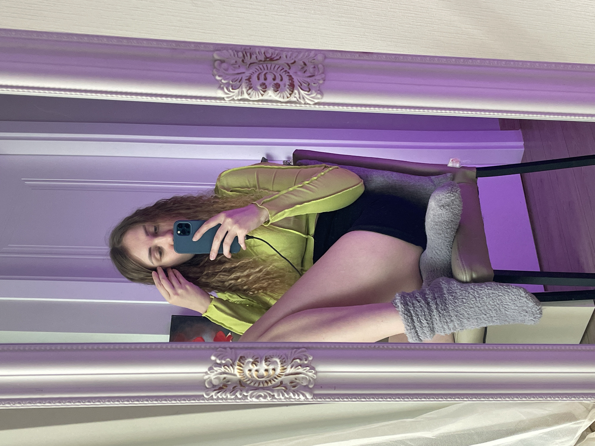 An indoor scene in Batumi, Georgia, captured as a mirror selfie. The image depicts a young adult female, seated on a grey upholstered chair, reflected in a large, ornate mirror with a decorative silver-white frame. She wears a lime green ribbed long-sleeve top with exposed stitching, black shorts, and grey fuzzy ankle socks. Her long, wavy, light brown hair is touched by her left hand, while her right hand holds a dark teal smartphone vertically, aiming it towards the mirror to take the photograph. A black cable is connected to the smartphone. Her eyes are directed downwards towards the phone screen. The background, visible in the mirror's reflection, consists of light-colored walls, some with vertical paneling, and a light wooden floor. The entire scene is strongly illuminated by a dominant purple ambient light source, and a framed piece of artwork with red elements is partially visible on the wall behind the subject's reflection.