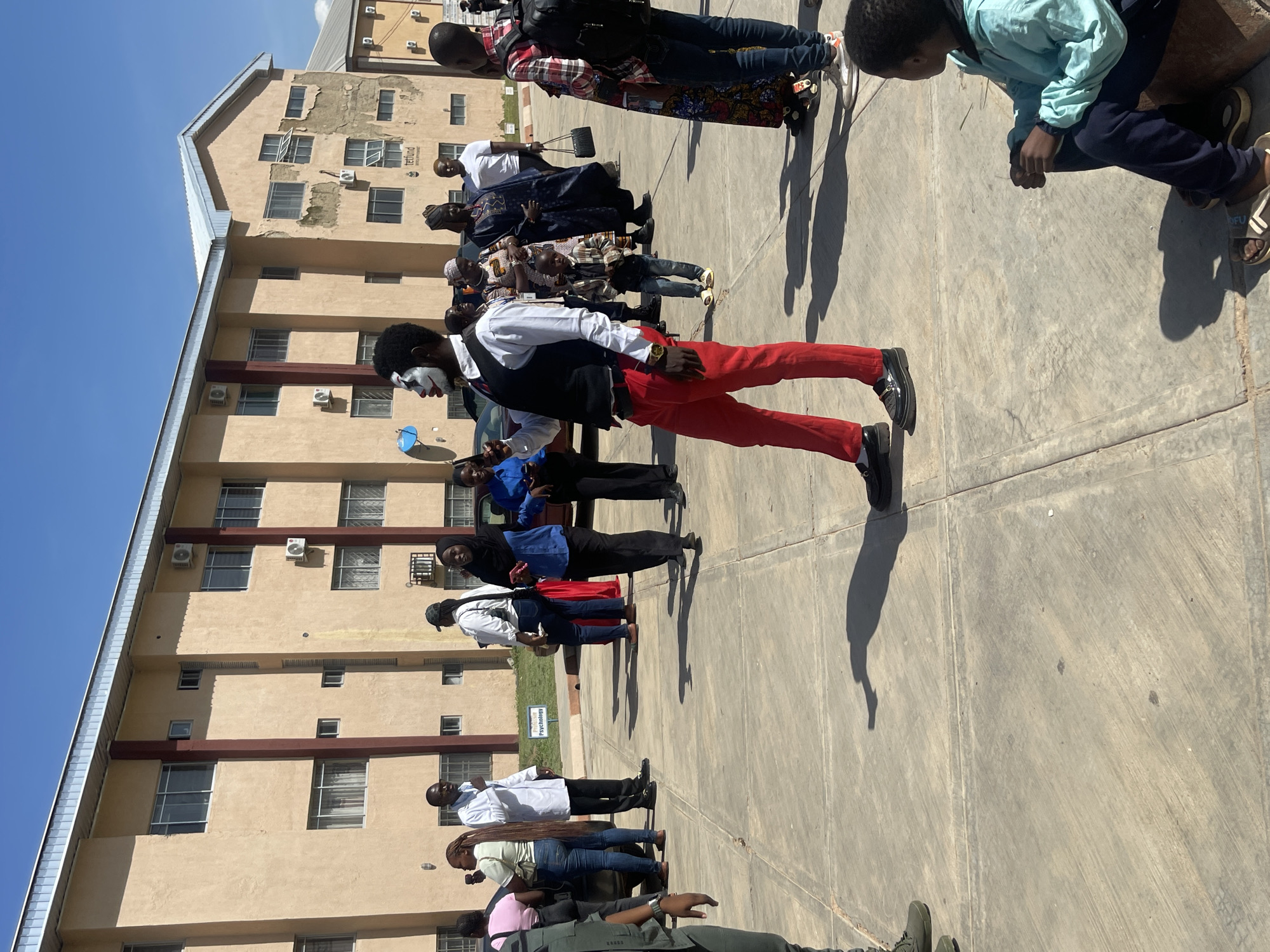 In Rafin Par, Nigeria, an outdoor scene unfolds on a paved area in front of a multi-story, tan-colored building. The building, with rows of windows and visible air conditioning units, appears to be a school or administrative complex. A group of people are gathered in the foreground and midground.
A man in the center foreground is dressed as a clown, with white face paint and dramatic eye makeup. He wears a black vest over a white shirt and bright red trousers, along with black dress shoes. He is the central figure and appears to be posing or performing.
To his left, a man in a white lab coat, likely a doctor or scientist, is speaking to others. Other individuals are seen in various states of interaction, including women in headscarves and traditional attire, and a young boy in a plaid shirt and jeans. The atmosphere appears to be a public gathering or event. The ground is concrete, marked with expansion joints, and a few shadows are cast by the people and the building. A sign indicating "Psychology" is visible in the background. The sky is clear and blue, suggesting a sunny day.