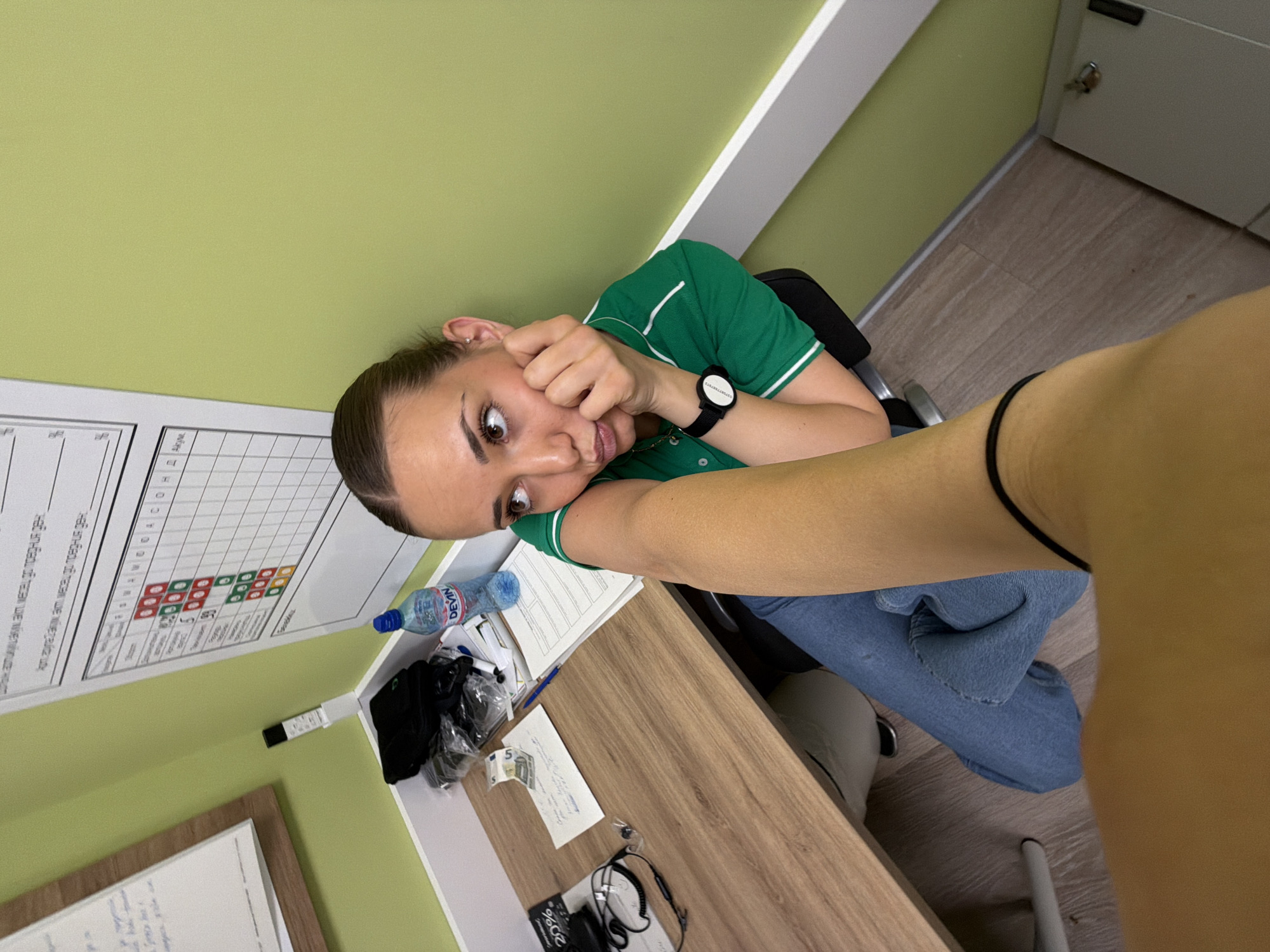 The image, taken indoors in Sofia, Bulgaria, depicts a young woman with brown hair and eyes, wearing a green polo shirt and jeans, seated at a wooden desk. She is resting her face on her hand, giving a wide-eyed expression directly to the camera. A black watch is visible on her wrist. On the desk are various items, including a water bottle labeled "Devin," black clothing or accessories, loose papers, a 5 Euro bill, and headphones. A whiteboard with charts and text is mounted on a green wall behind the desk. Part of another person's arm is visible in the foreground. A cabinet is seen to the right of the desk.