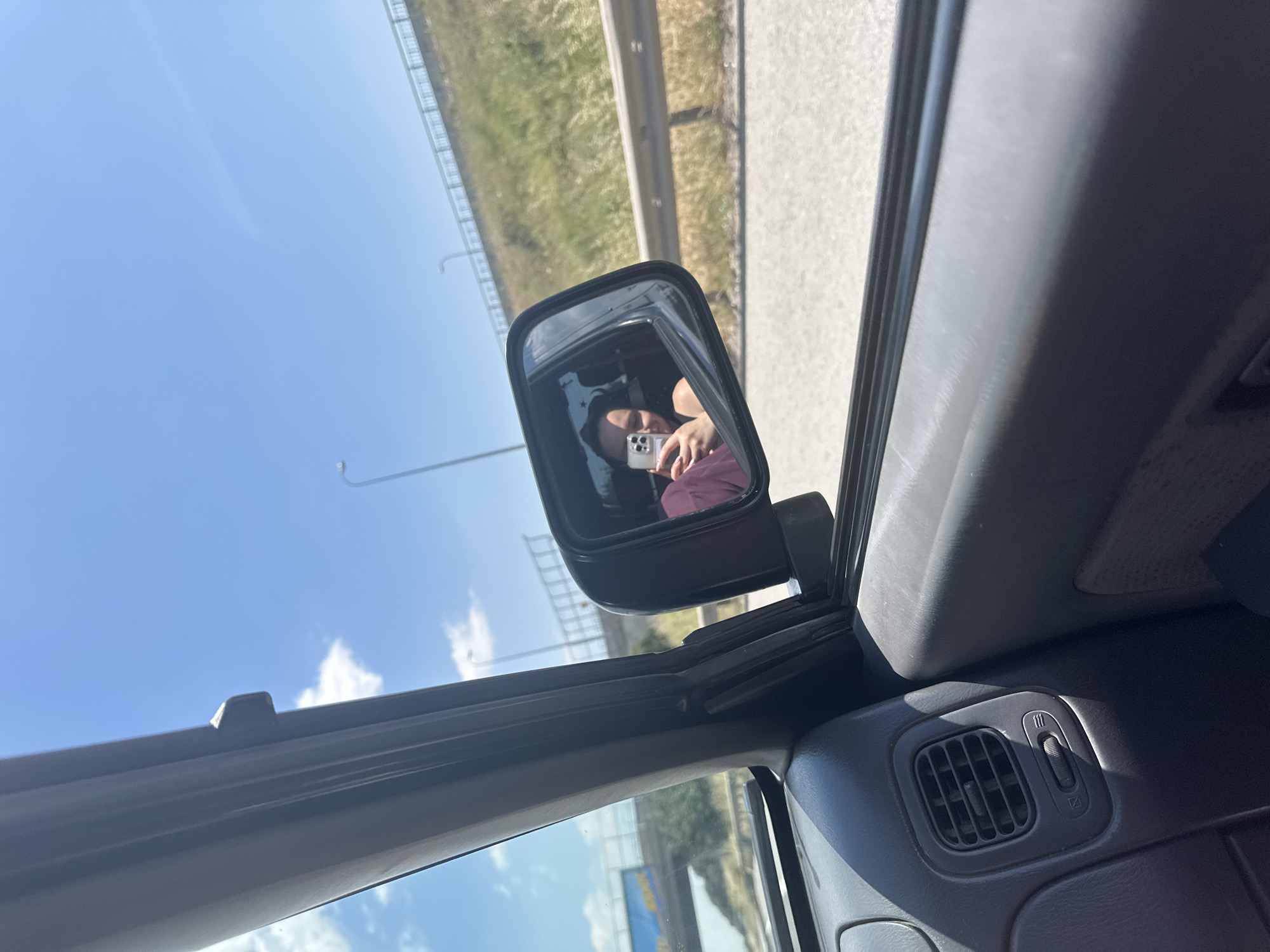 The image shows a view from inside a car, likely the driver's seat. In the side mirror, a person is visible holding a mobile phone, possibly taking a photo. The scenery reflected in the mirror appears to be a highway with a grassy shoulder and a blue sky. The interior of the car is visible, including the dashboard with an air vent and a control switch. Based on the context provided in the prompt, the car is likely located in or near Tuzla, Turkey, given the location information.