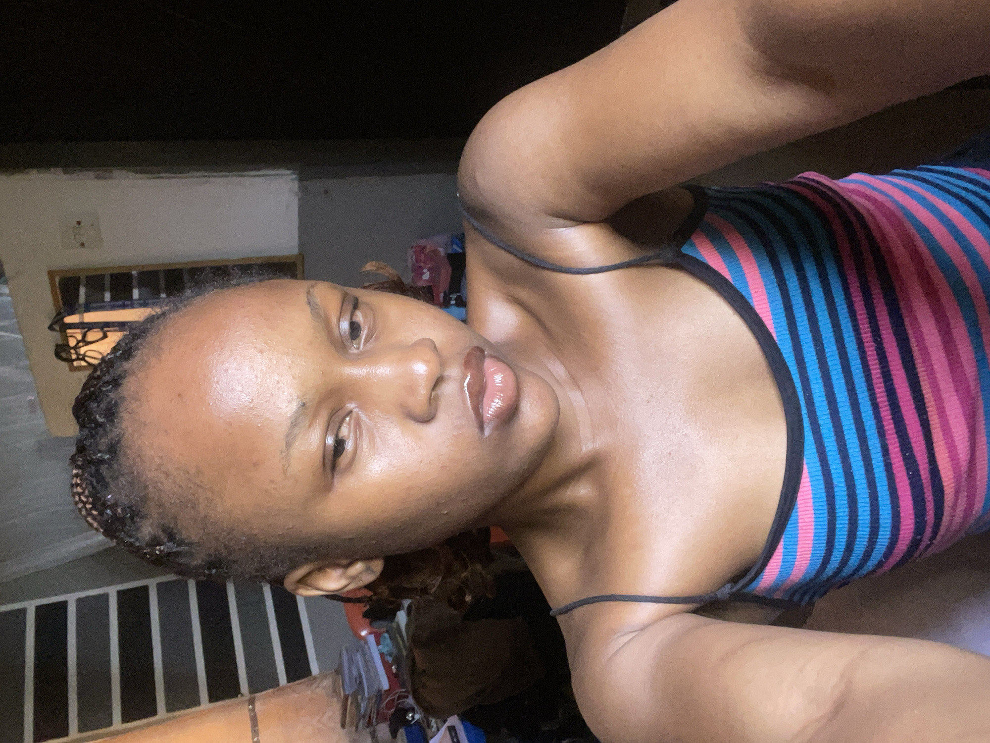 A young woman with braided hair and a pink, blue, and black striped tank top is taking a selfie. She is looking to the side with a slightly downward gaze. Her skin appears slightly oily. Behind her, a wall is visible with some objects on it. To her left, a stacked shelf unit with books and other items is partially obscured. The environment appears to be indoors, possibly a bedroom or living area. The lighting is soft, suggesting it is daytime. There is no specific indication of the location in Jos, Nigeria, beyond it being an indoor residential setting.