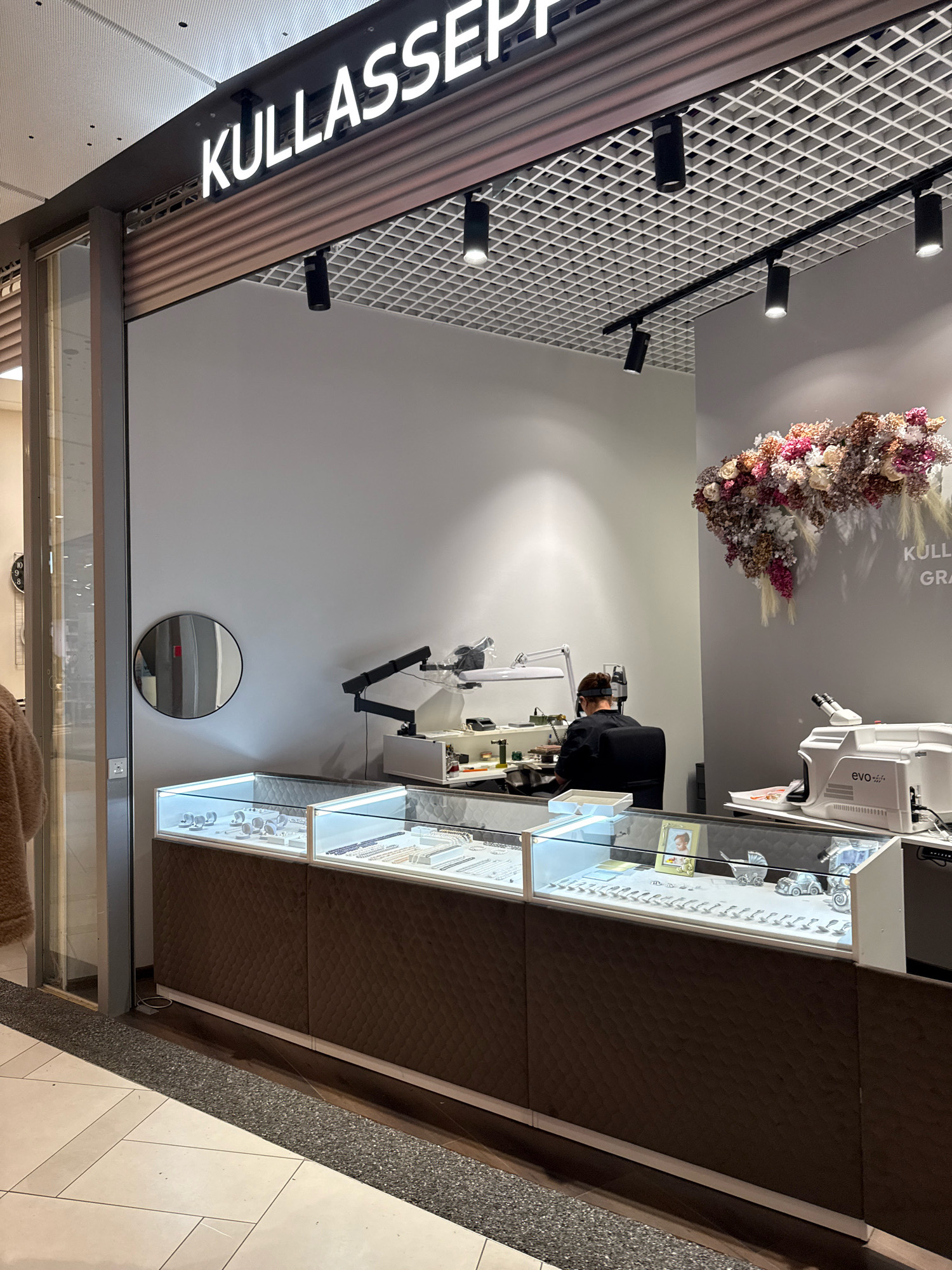 The image shows the exterior of a jewelry store named "KULLASSEPP" in Tallinn, Estonia. The store has a clean, modern facade with a prominent illuminated sign. Inside, display cases showcase various jewelry items, and a person is seated at a workbench, appearing to be working on or examining jewelry under a magnification lamp. A microscope is also visible on a counter. The store is well-lit, and the overall impression is one of a professional establishment. The flooring is tiled in a light color.