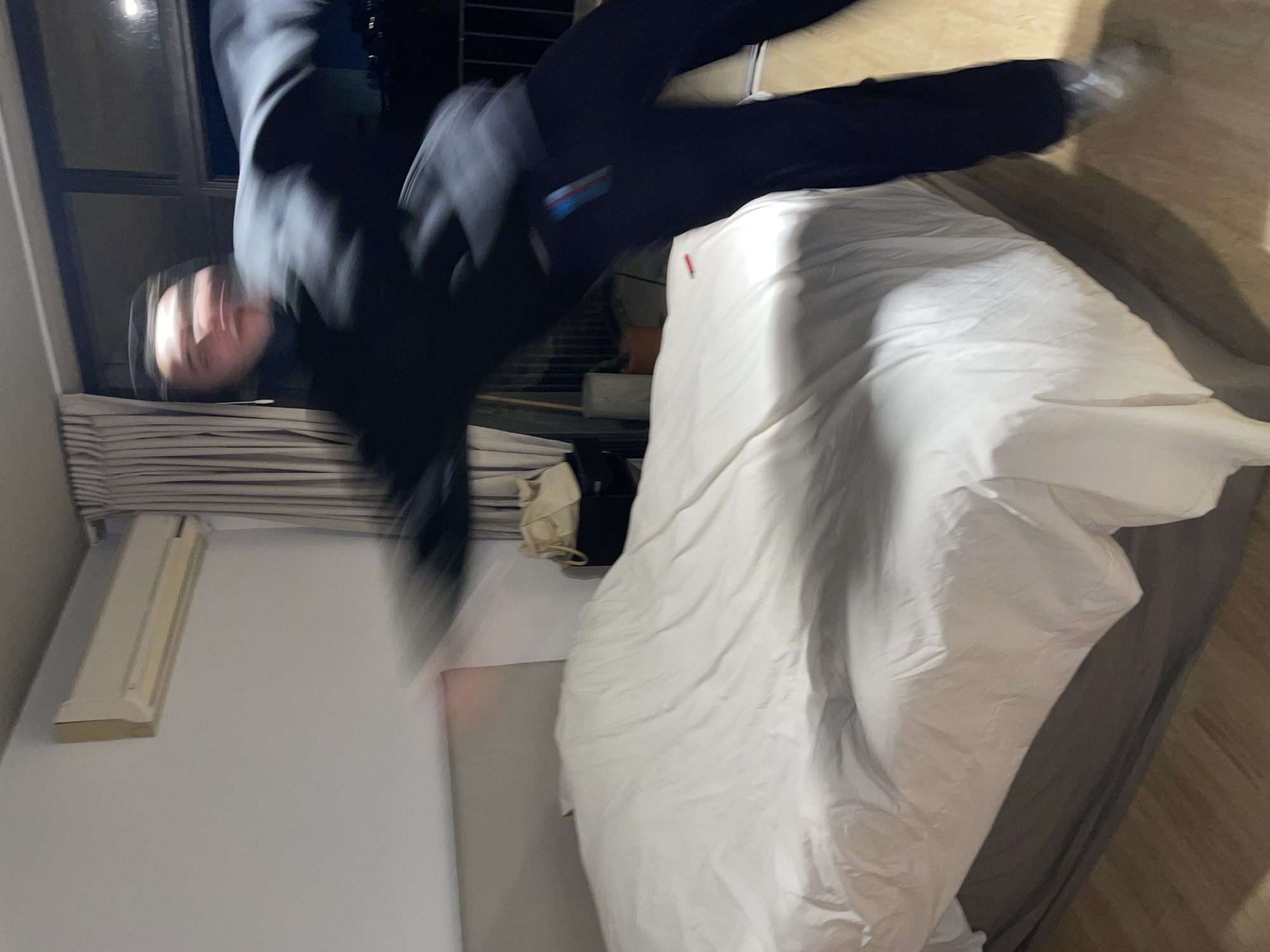 A person wearing dark clothing is mid-air, above a white-covered bed in a room. The room contains a window, blinds, a wall-mounted air conditioning unit, and flooring with wood grain. The location is not discernible from the image.