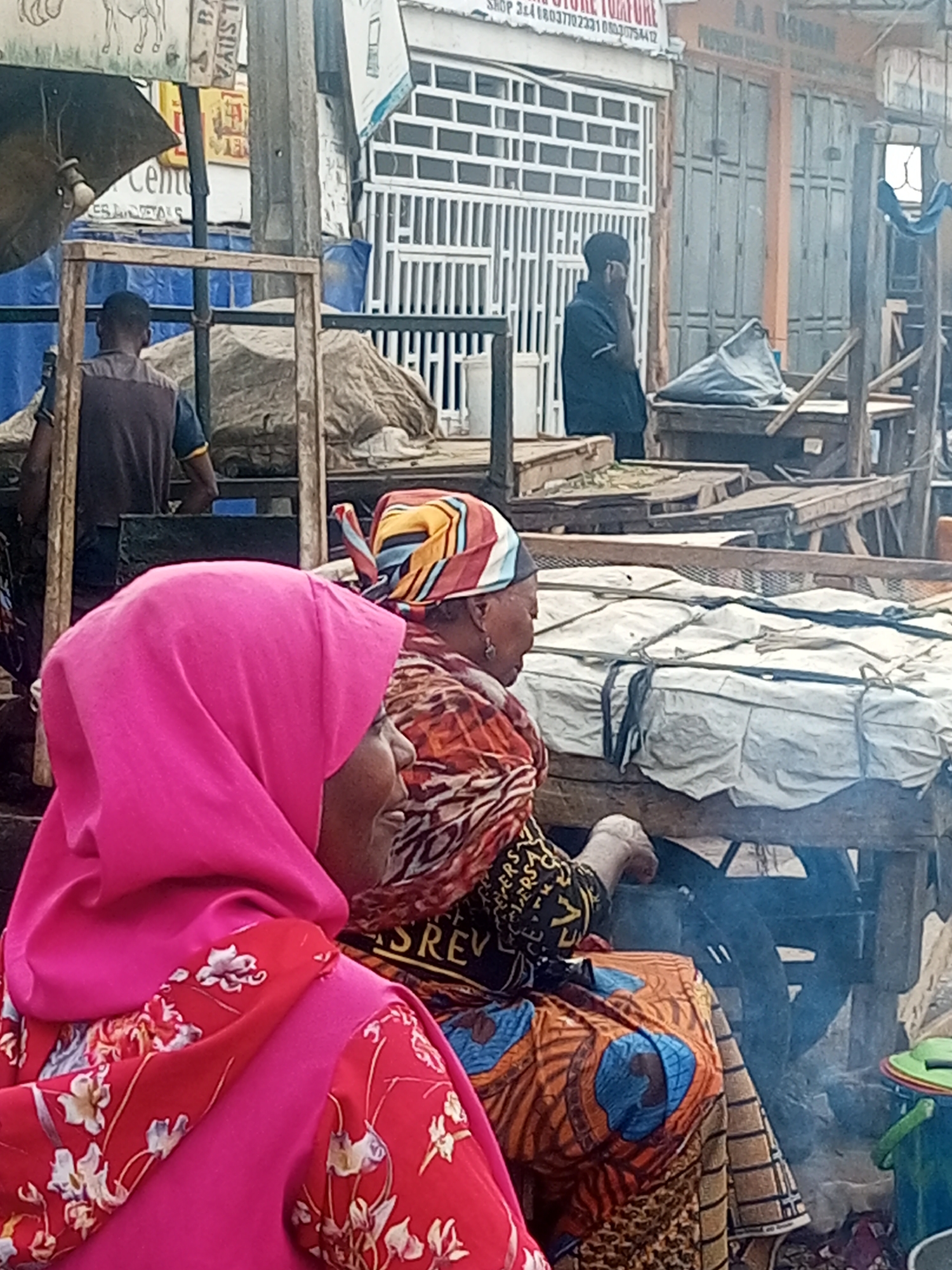Two women are in the foreground. One wears a bright pink headscarf with a red floral print. The other wears a multicolored headscarf and a patterned orange dress. Both women are sitting near a table with a white tarp tied down. Smoke rises in front of the table. In the background, a man in a dark shirt is standing near a stall, appearing to be on the phone. Other stalls and structures are visible, with signs indicating businesses like "AA Usman Provision Store" and "Shop 3&4." The scene is bustling, suggesting a marketplace setting in Tempure, Nigeria.