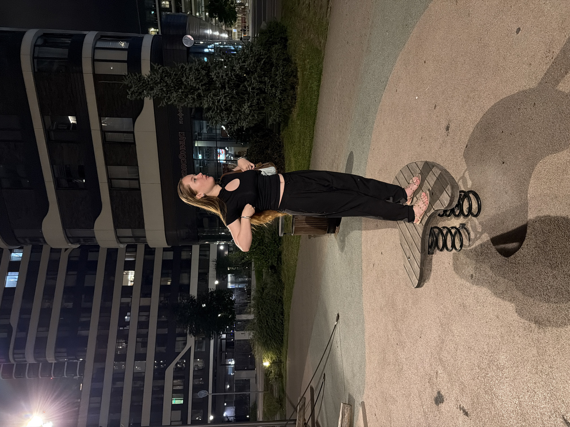 A young woman with long blonde hair stands on a spring-mounted playground toy at night in Moscow, Russia. She is wearing a black crop top and black wide-leg pants, and gold studded sandals. She holds her hands up to the sides of her torso, adjusting her clothing. A small purse is slung over her right shoulder. In the background, a modern, multi-story building with curved balconies and illuminated windows dominates the scene. A dark evergreen tree stands to the right of the building. The ground of the playground is a textured, speckled surface in shades of beige and grey. A trash can is visible to the left of the woman. The lighting suggests artificial streetlights illuminating the area.