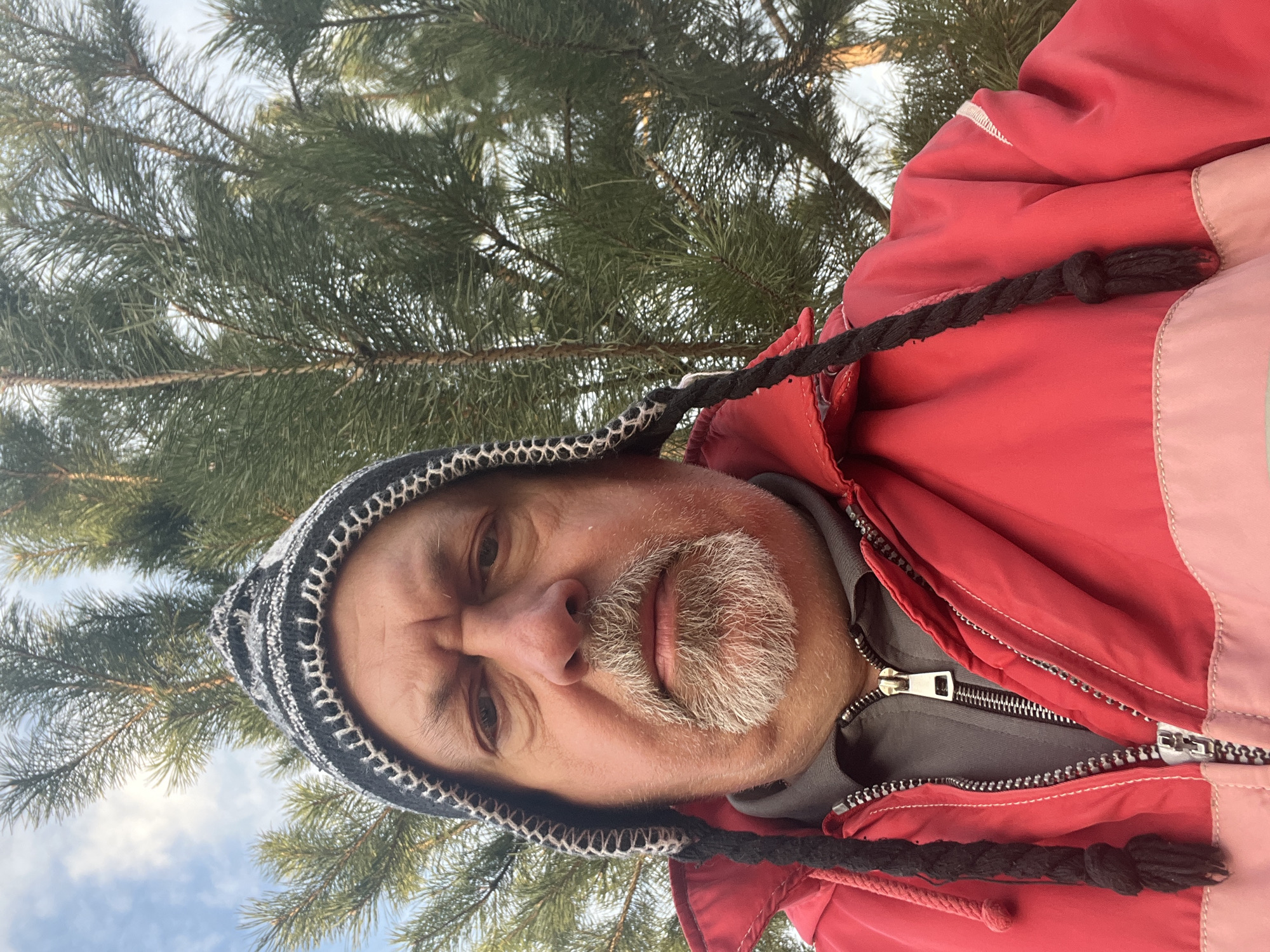 A low-angle close-up shot shows a man in a hooded hat and red jacket with tree branches in the background, possibly located near Podolsk, Russia. The man has a salt-and-pepper beard and a stern expression. He is wearing a black and white patterned hat with black braided drawstrings. The jacket is red with a visible zipper. The background features green pine tree branches and a glimpse of a blue sky with white clouds.