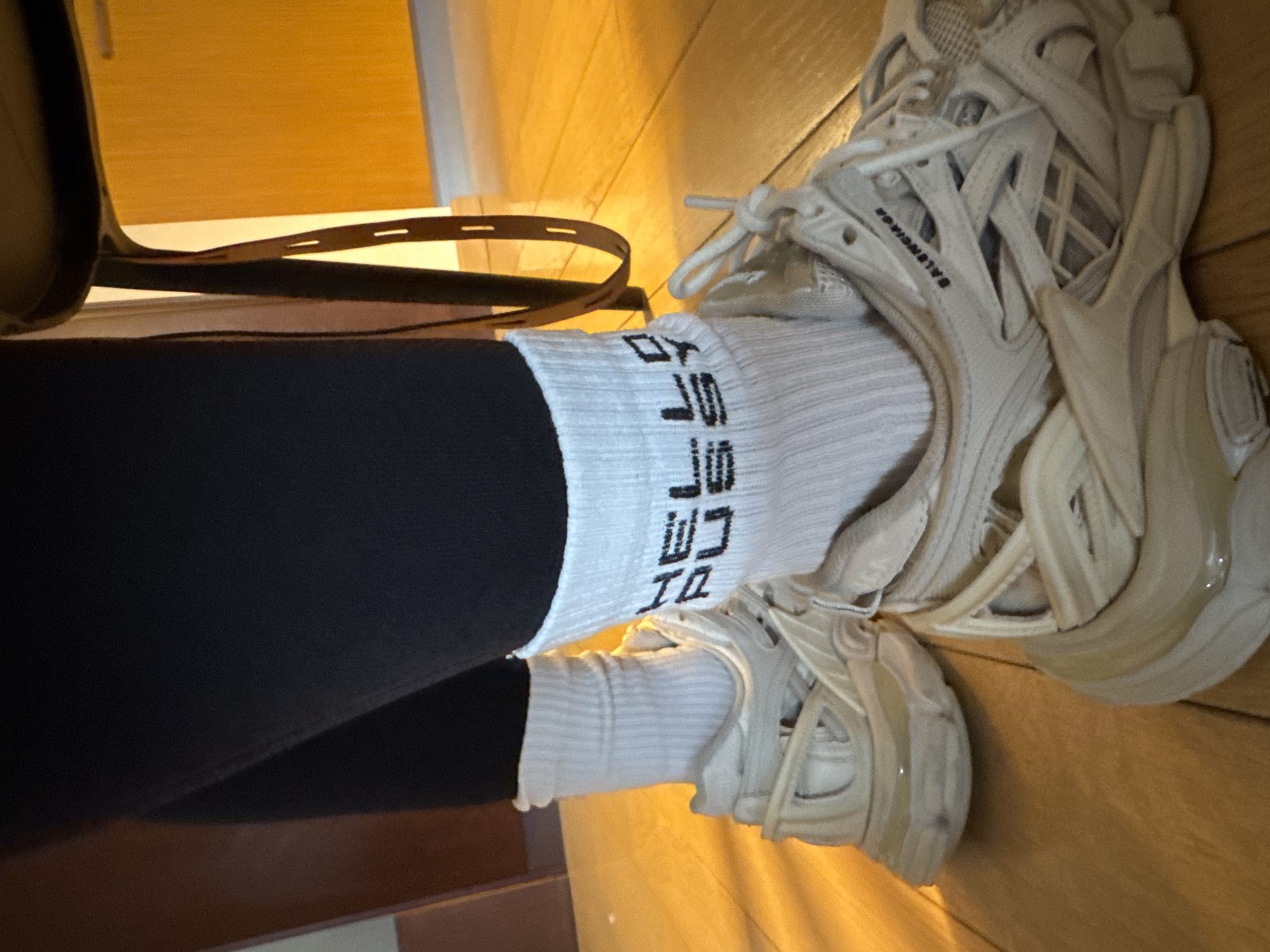 A close-up shot of a person's legs and feet in a possible domestic setting, the person wears black leggings tucked into white ribbed socks with black lettering, and cream-colored Balenciaga sneakers. The background includes what seems to be a wooden floor and a yellow-toned wall or cabinet, along with a glimpse of what might be a bag strap. The location is Unknown, Unknown.