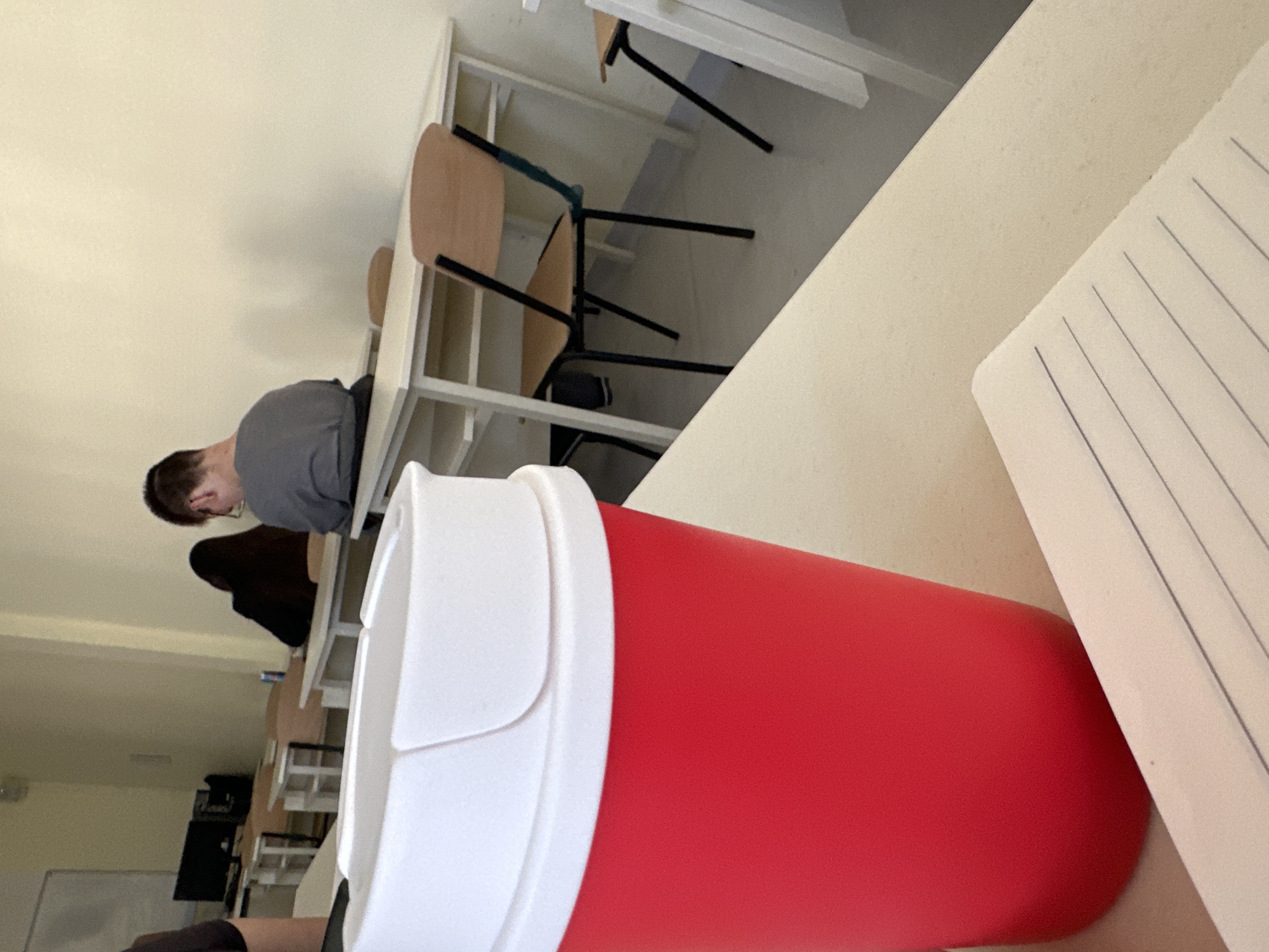 A red and white tumbler sits on a light-colored desk with lined paper. The image is taken in a classroom setting, possibly a university, in Sofia, Bulgaria. A person with short brown hair and wearing a gray shirt is seated at a desk in the background, along with other students and desks arranged in rows. A light wall is visible, and a window is on the left.