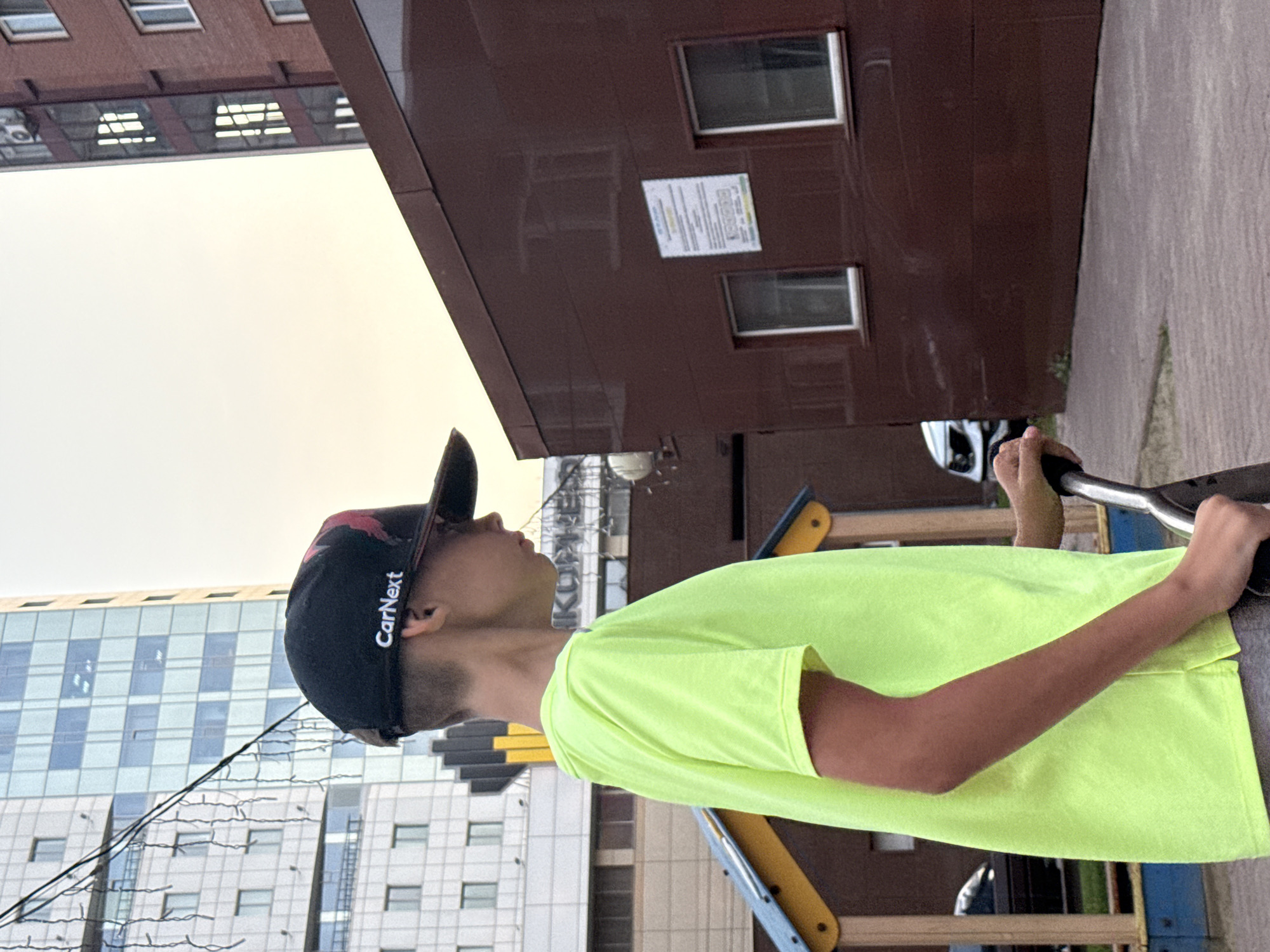 A young person wearing a neon yellow t-shirt and a black baseball cap is standing outside, seemingly looking up at a building. The cap has the word "CarNext" visible on the side. The background features a modern, multi-story building with a reddish-brown exterior and numerous windows. Further in the background, another tall building with a glass facade and a grid-like pattern of windows is visible. The scene appears to be an urban setting in Krasnoyarsk, Russia. The lighting suggests it is daytime, possibly overcast, as there are no strong shadows. The ground beneath the person is paved. The person's gaze is directed upwards, and their posture suggests they are perhaps waiting or observing something. The overall mood is neutral.