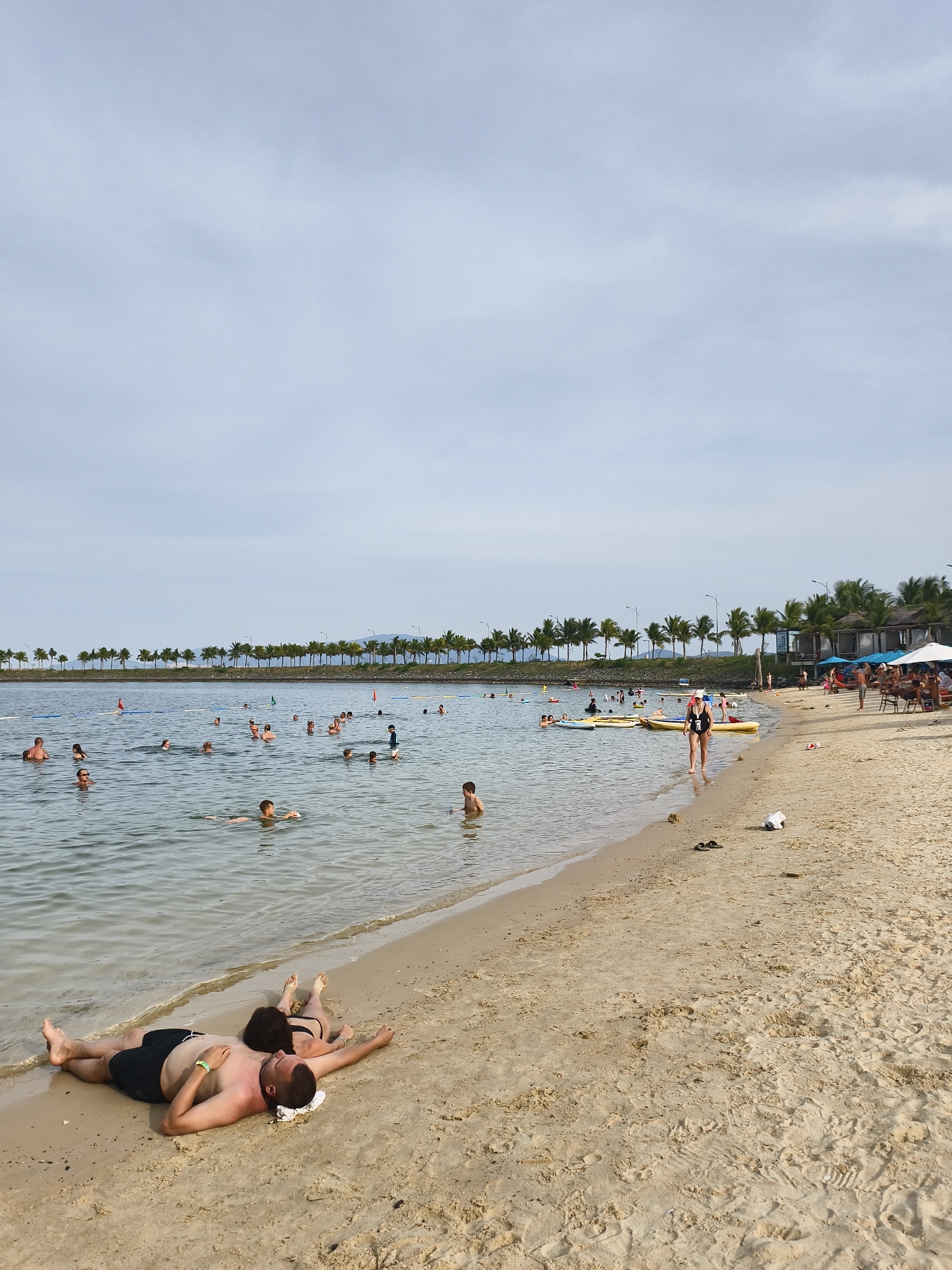 A wide shot captures a beach scene in Bac Nha Trang Ward, Vietnam. The foreground features a sandy beach with two individuals lying face down in the sand near the water's edge. The male is wearing black swim shorts, and the female is in a black bikini. To the left, the ocean is populated with numerous people swimming and wading. Several yellow inflatable rafts are visible in the water. The middle ground is dominated by a line of palm trees bordering the beach, with some resort-like structures partially obscured by the trees. The background shows a hazy sky and distant mountains. The overall impression is a popular, sunny day at a beach resort.