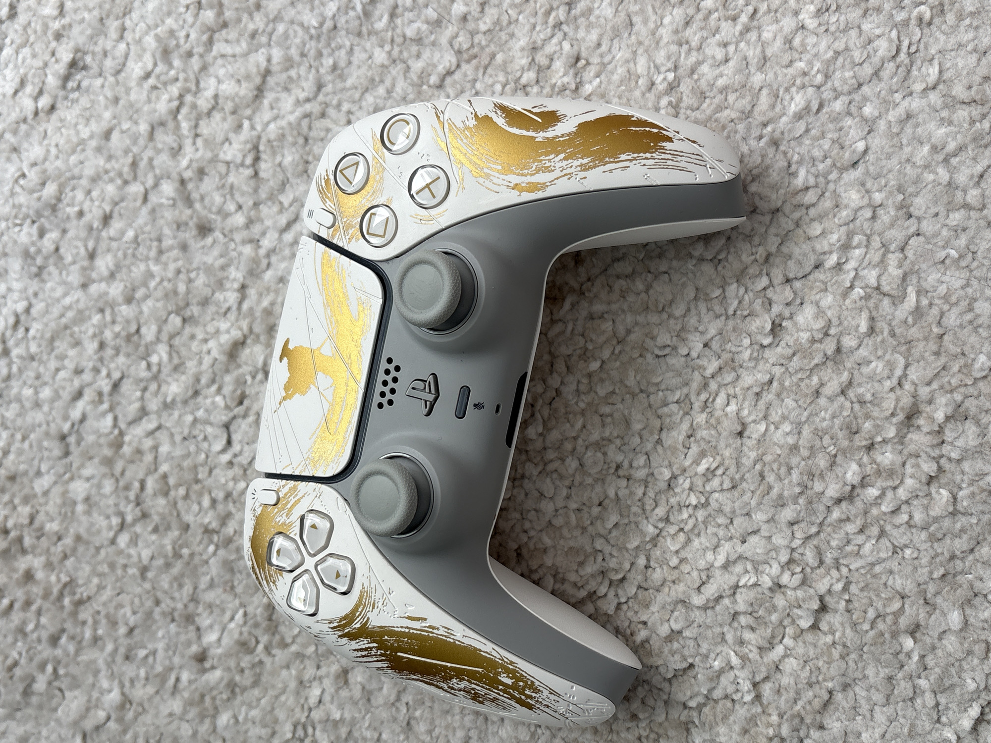 A custom-designed PlayStation 5 DualSense wireless controller rests on a light gray, shaggy carpet. The controller is predominantly white with a distinct gold brushstroke and linear design across its faceplate and grip areas. The central faceplate and main body panels are white, while the analog sticks and inner grips are a uniform medium gray. The D-pad and action buttons (triangle, circle, cross, square) are translucent with gold symbols, corresponding with the gold aesthetic. The PlayStation logo button is black. The carpet features a high pile and an irregular texture, showing variations in fiber density and minor shadows cast by the controller. A few fine, dark fibers are visible on the carpet's surface in the lower right area of the primary image and some cropped sections. The lighting is even, suggesting an indoor setting. There are no people or other objects present in the scene.