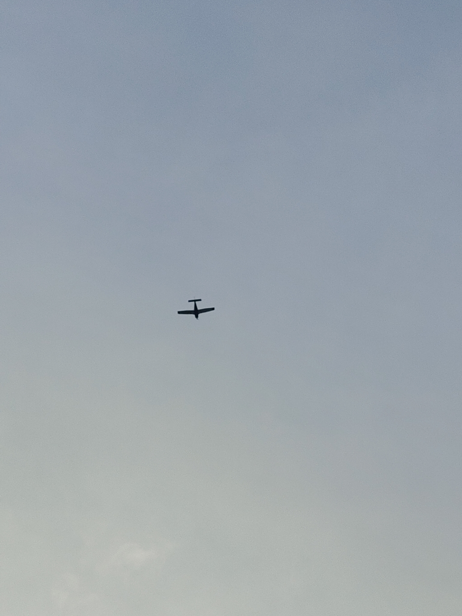 This media file shows a small airplane in silhouette against a pale, overcast sky. The airplane has a single propeller and appears to be a light aircraft. It is positioned in the upper-middle portion of the frame. The sky is a uniform, muted blue-grey, suggesting it might be late afternoon or early morning, or possibly a day with significant cloud cover. There are no discernible landmarks or location cues within the frame, beyond the implied open sky. The image is still and there is no indication of any activity or event beyond the plane's presence in the sky. There is no visible text in the image. The context of Nowy Dwór Mazowiecki, Poland, suggests the plane is likely flying over or near this area.