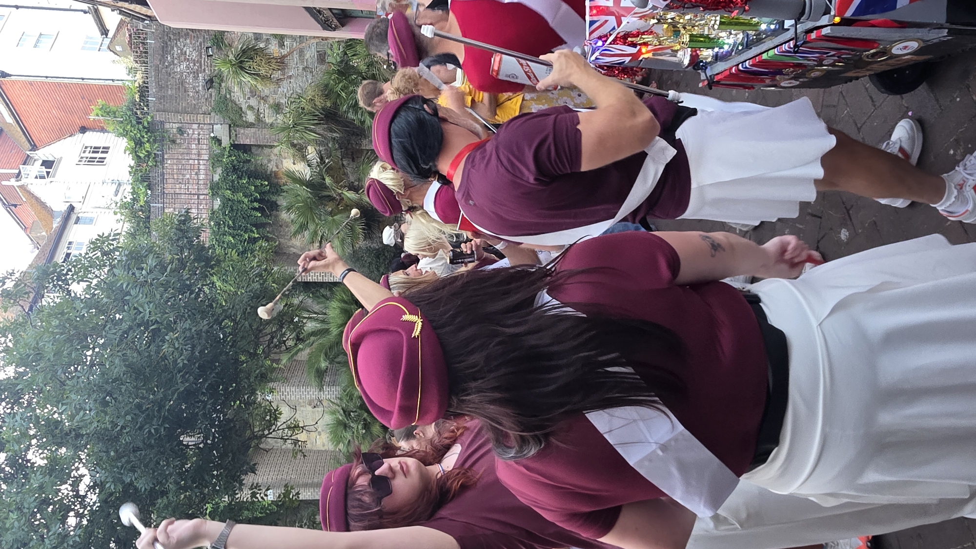 In St Leonards, United Kingdom, a group of individuals in matching maroon uniforms with white sashes participate in an outdoor event. The uniforms consist of maroon tops and white skirts or trousers, with matching maroon caps adorned with gold trim and a wheat stalk emblem. Some individuals are holding drumsticks, suggesting a parade or performance. One person, seen from behind, wears a black belt over their maroon top and holds a red and silver can. Another person, partially visible on the left, wears sunglasses and a maroon cap. The background features brick walls, dense green foliage, palm trees, and buildings with tiled roofs. In the foreground, a cart is decorated with numerous trophies and medals, some featuring Union Jack motifs, indicating a celebratory or competitive event. The lighting suggests it is daytime.