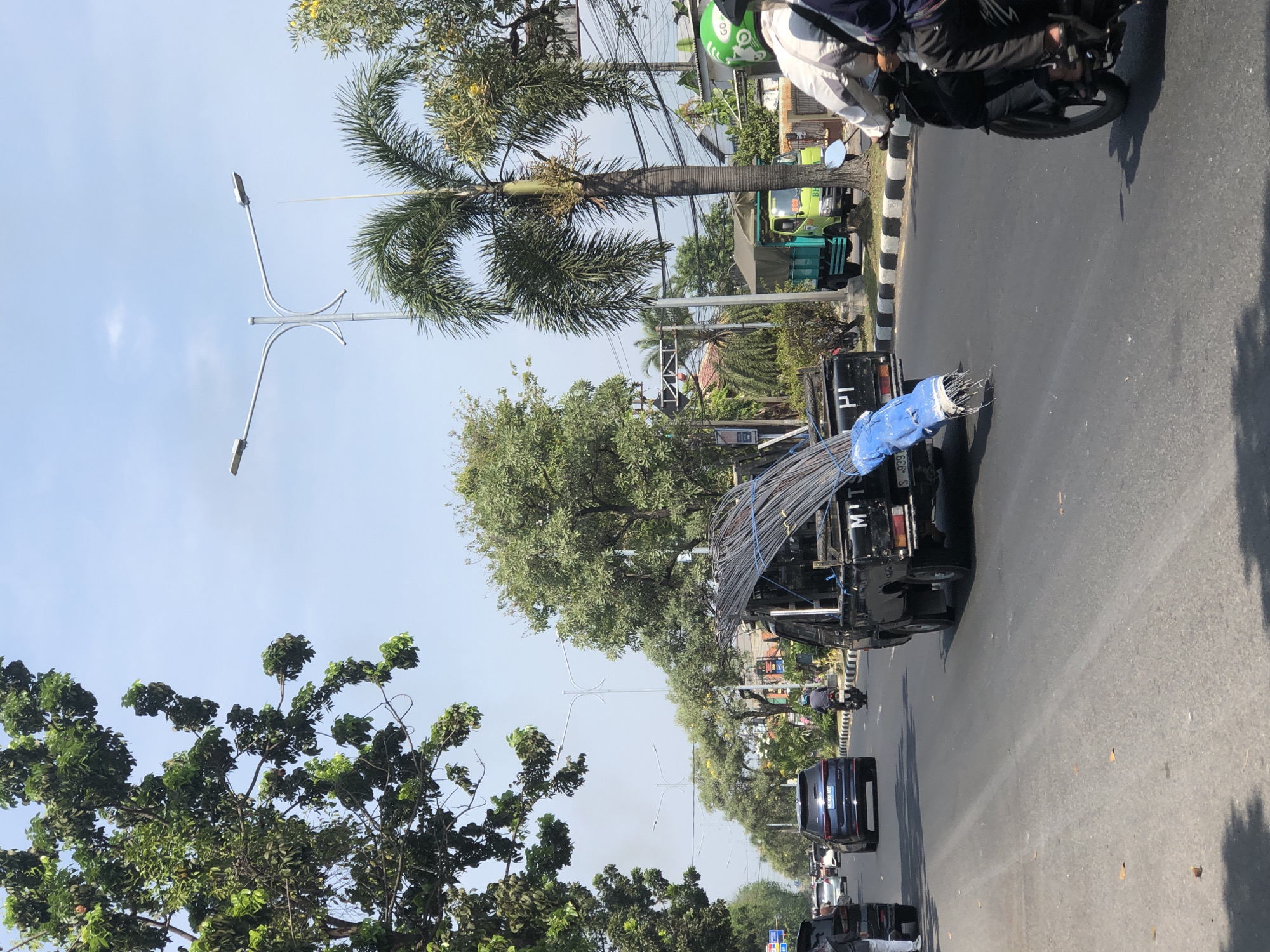 In Larangan, Indonesia, a daytime scene unfolds on a multi-lane asphalt road under a clear sky. The road is lined with trees and streetlights, with some buildings and greenery visible in the background. Traffic consists of various vehicles, including cars, a green truck, a motorcycle with two occupants, and a black pickup truck. The pickup truck is heavily laden with a bundle of metal rods, secured with a blue tarp. The metal rods are the primary focus, extending prominently from the rear of the truck. One person is clearly visible on the motorcycle, wearing a helmet.